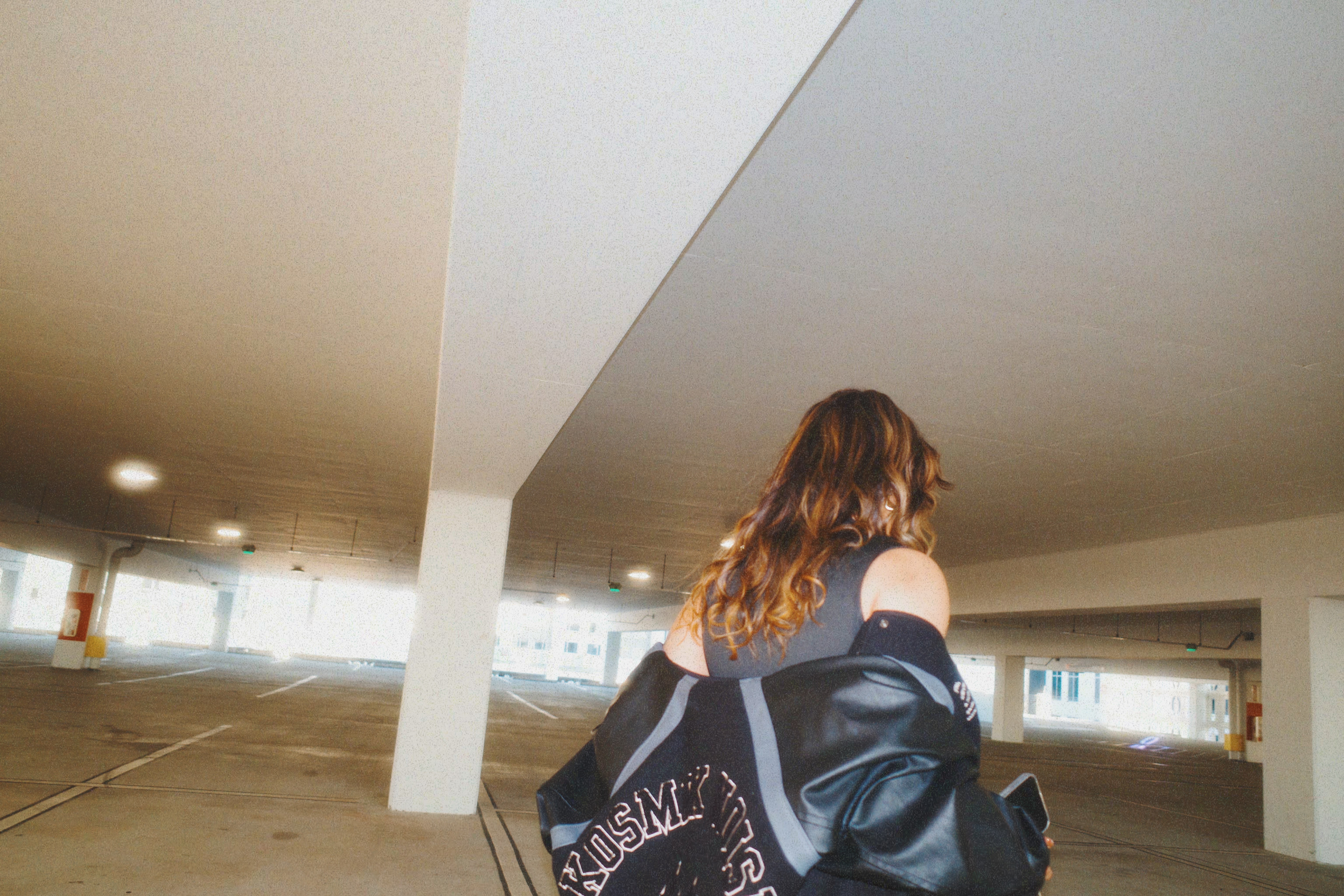 Person with shoulder-length hair standing in a parking garage, facing away, wearing a dark jacket and carrying a bag.