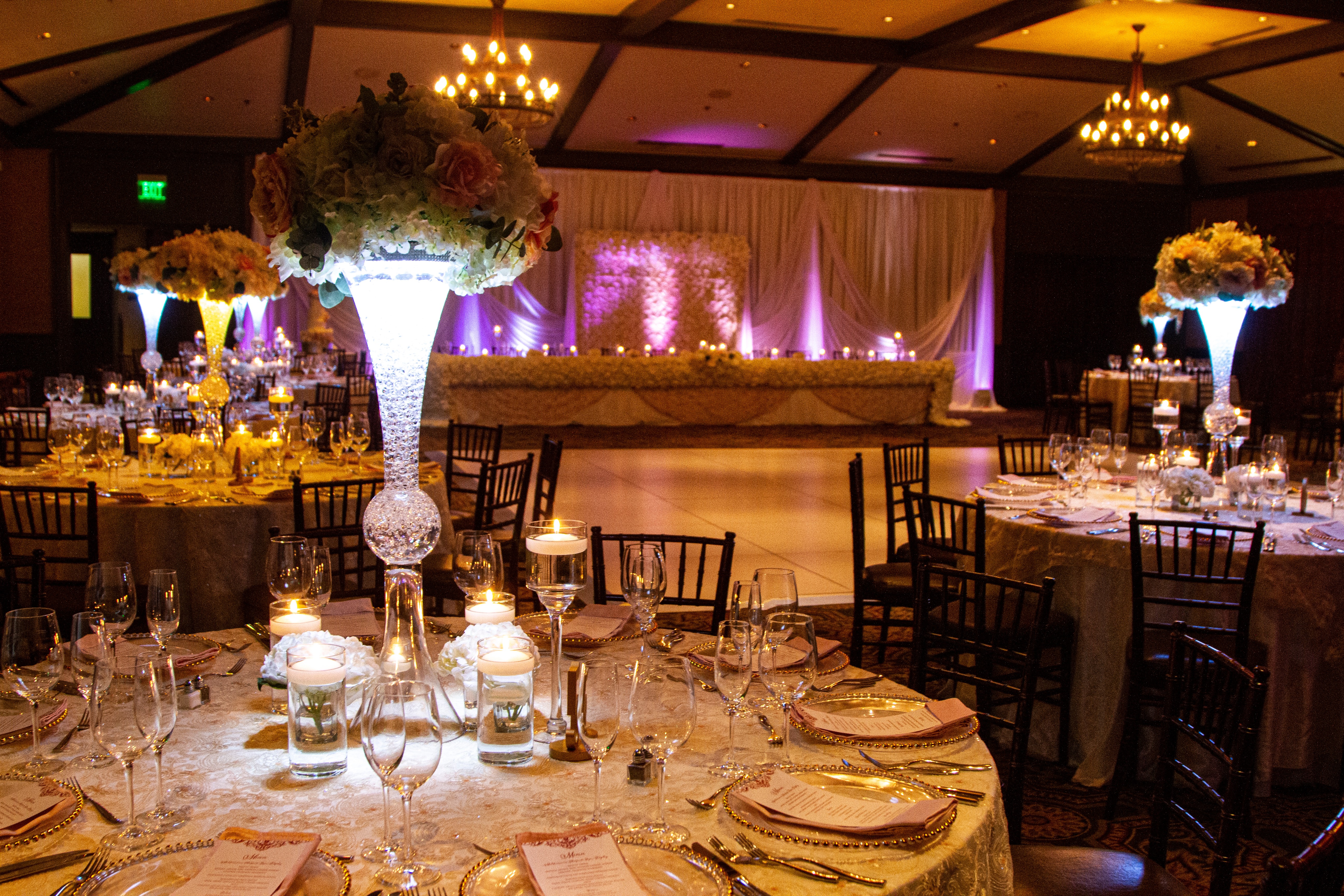 Elegant banquet hall with floral centerpieces, table settings, and draped curtains, illuminated by chandeliers and purple lighting.