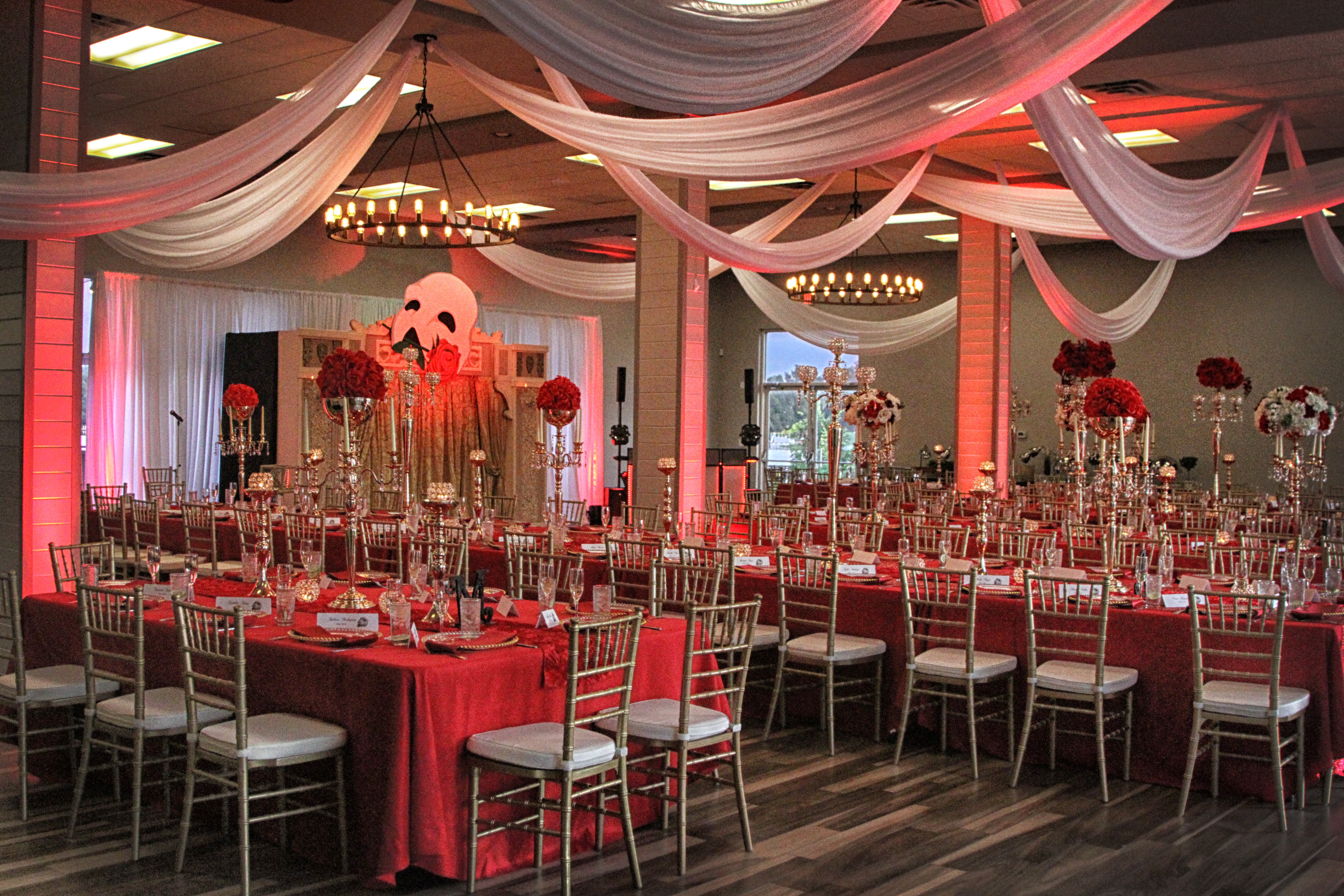 Elegant event space decorated with draped fabric, floral centerpieces, and red accents, with tables set for a celebration.