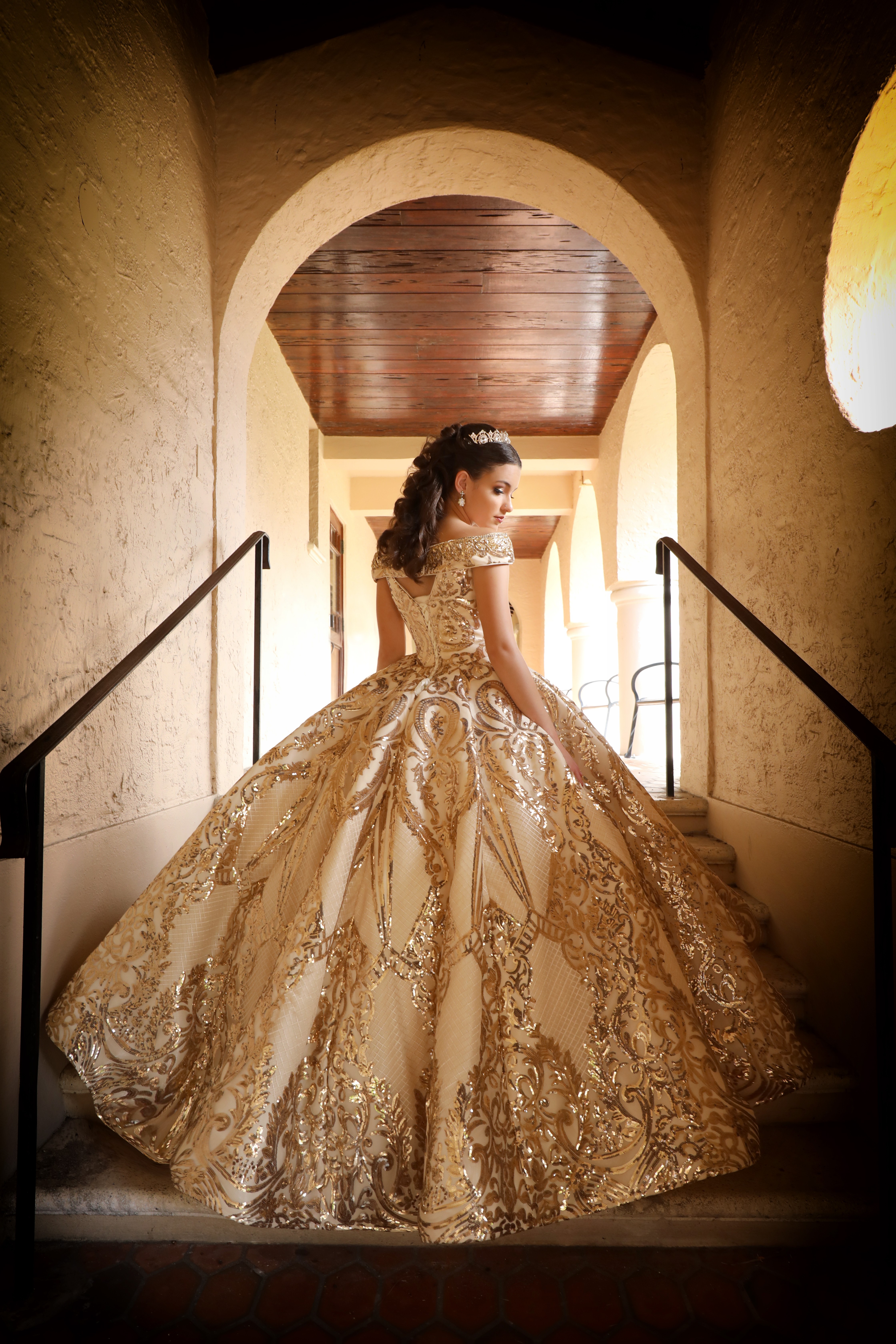 Person in an ornate gold and cream ball gown with a full skirt, standing in a hallway with arches, facing away.