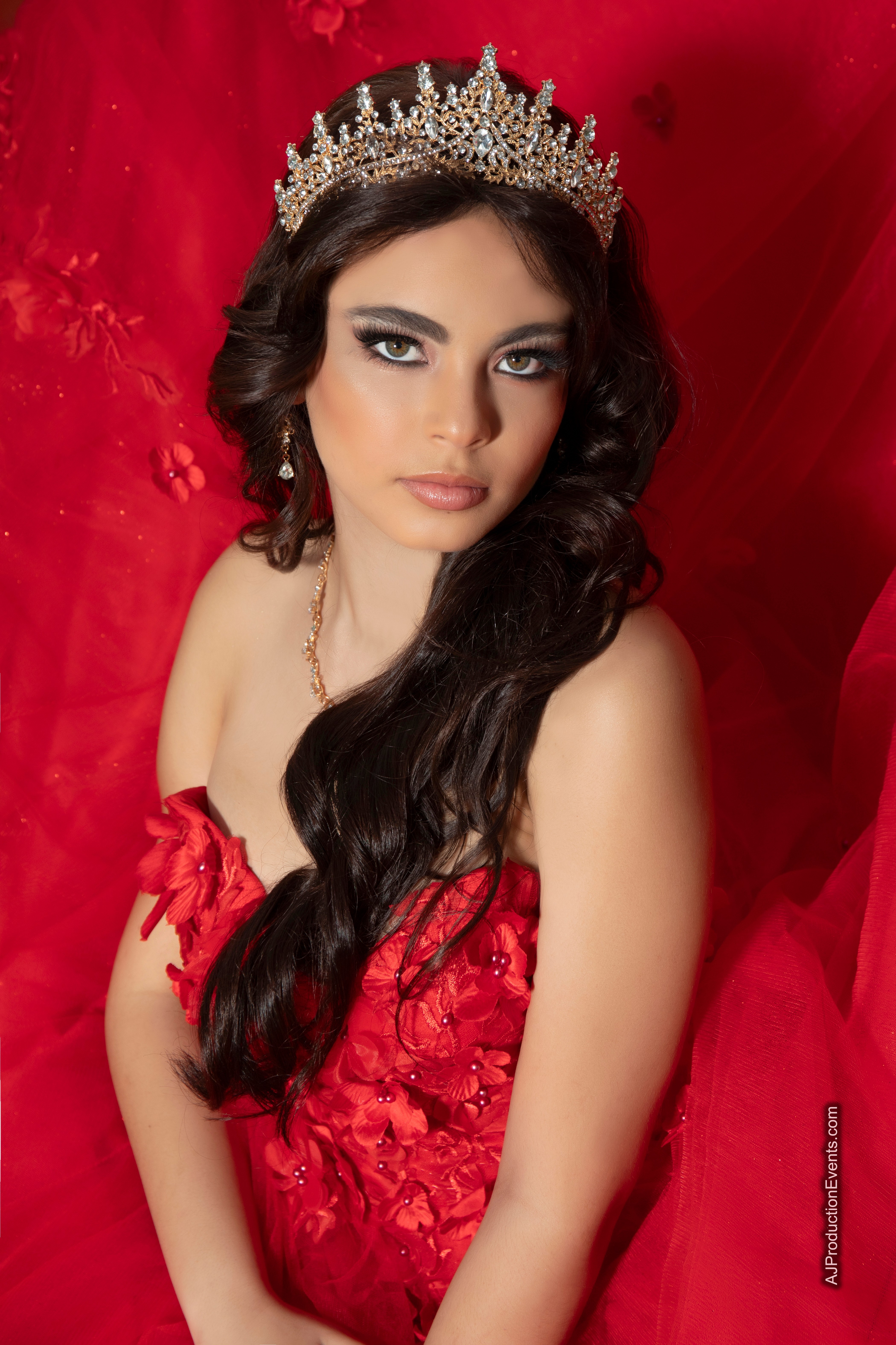 Young woman with dark hair wearing a crown and red dress, posed against a red background.