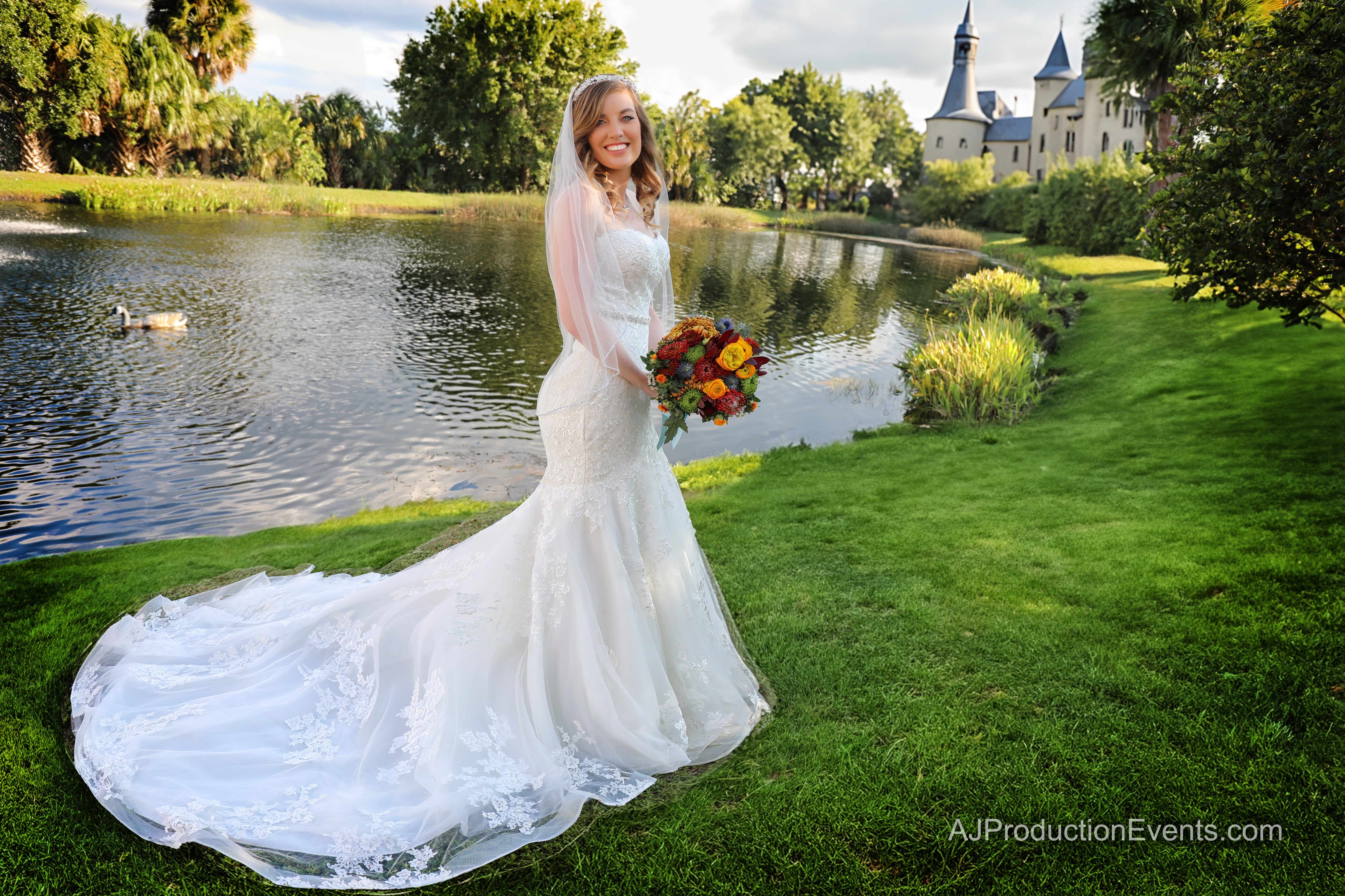 Bride in a white wedding dress holding a bouquet, standing on grass near a pond with a castle in the background.