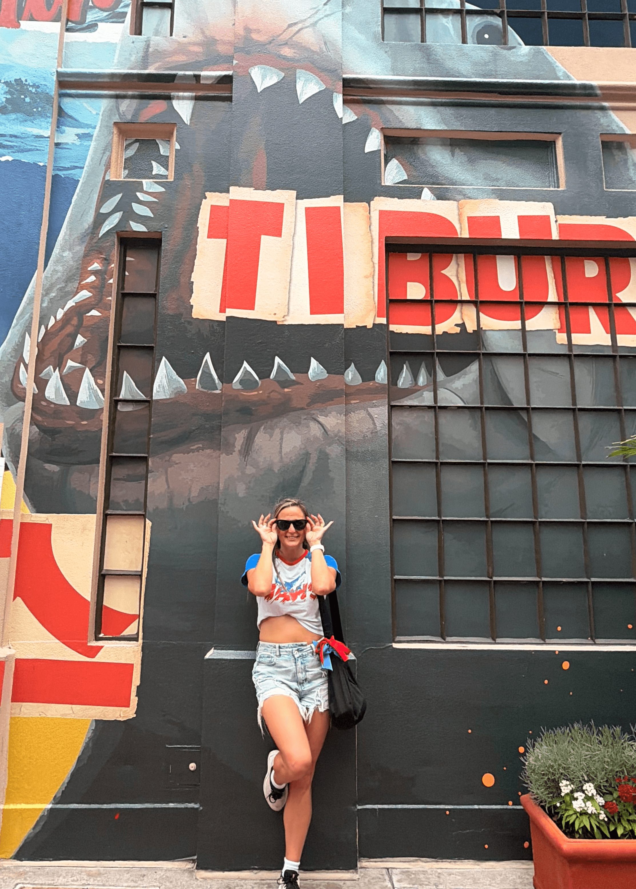 Person standing in front of a large mural on a building, wearing sunglasses, a crop top, and shorts, with hands raised.