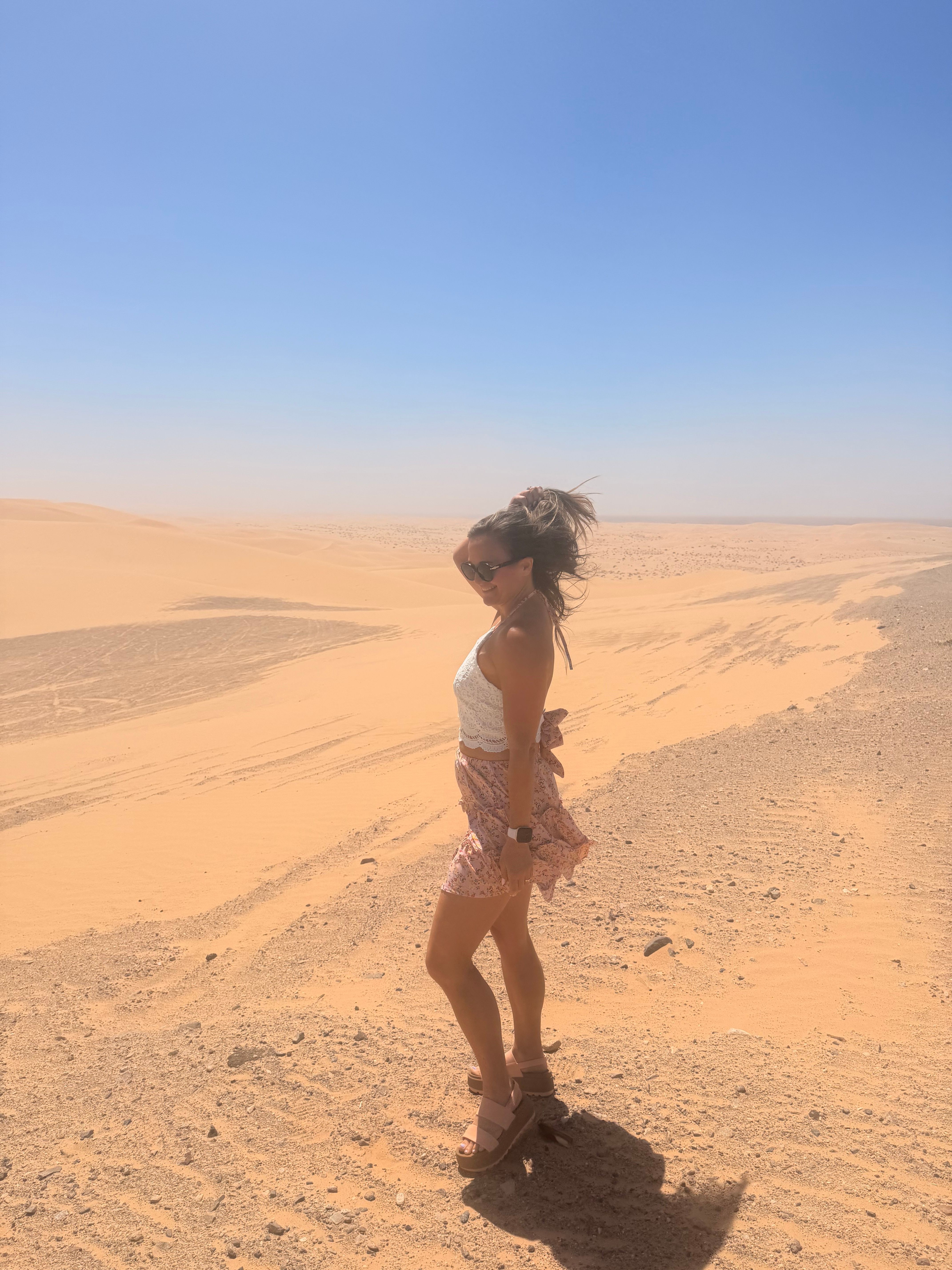 Person walking on sandy desert with clear blue sky, facing sideways, shadow visible on ground.