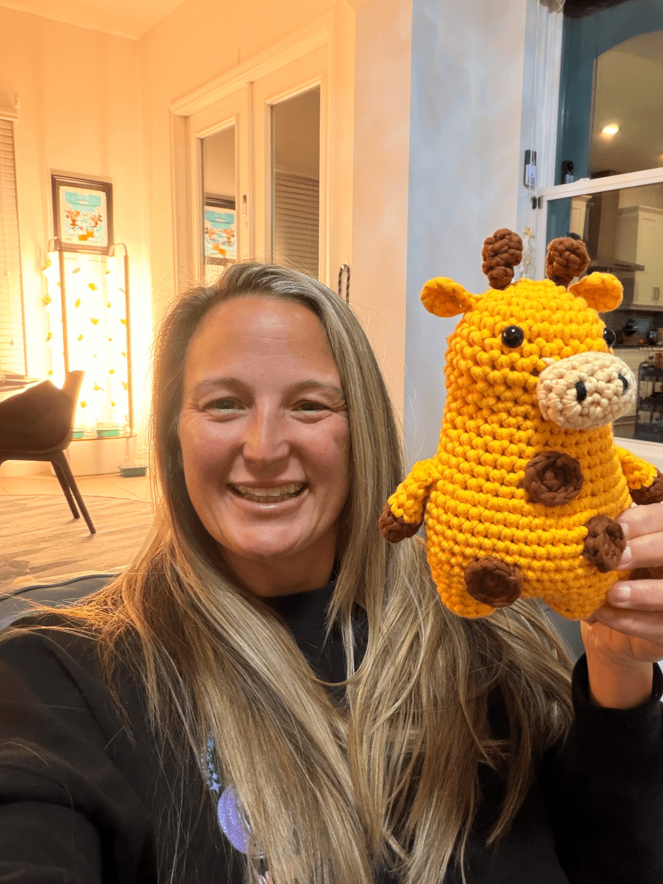 Woman smiling holding a yellow crocheted giraffe plush toy in a cozy room with windows and artwork.