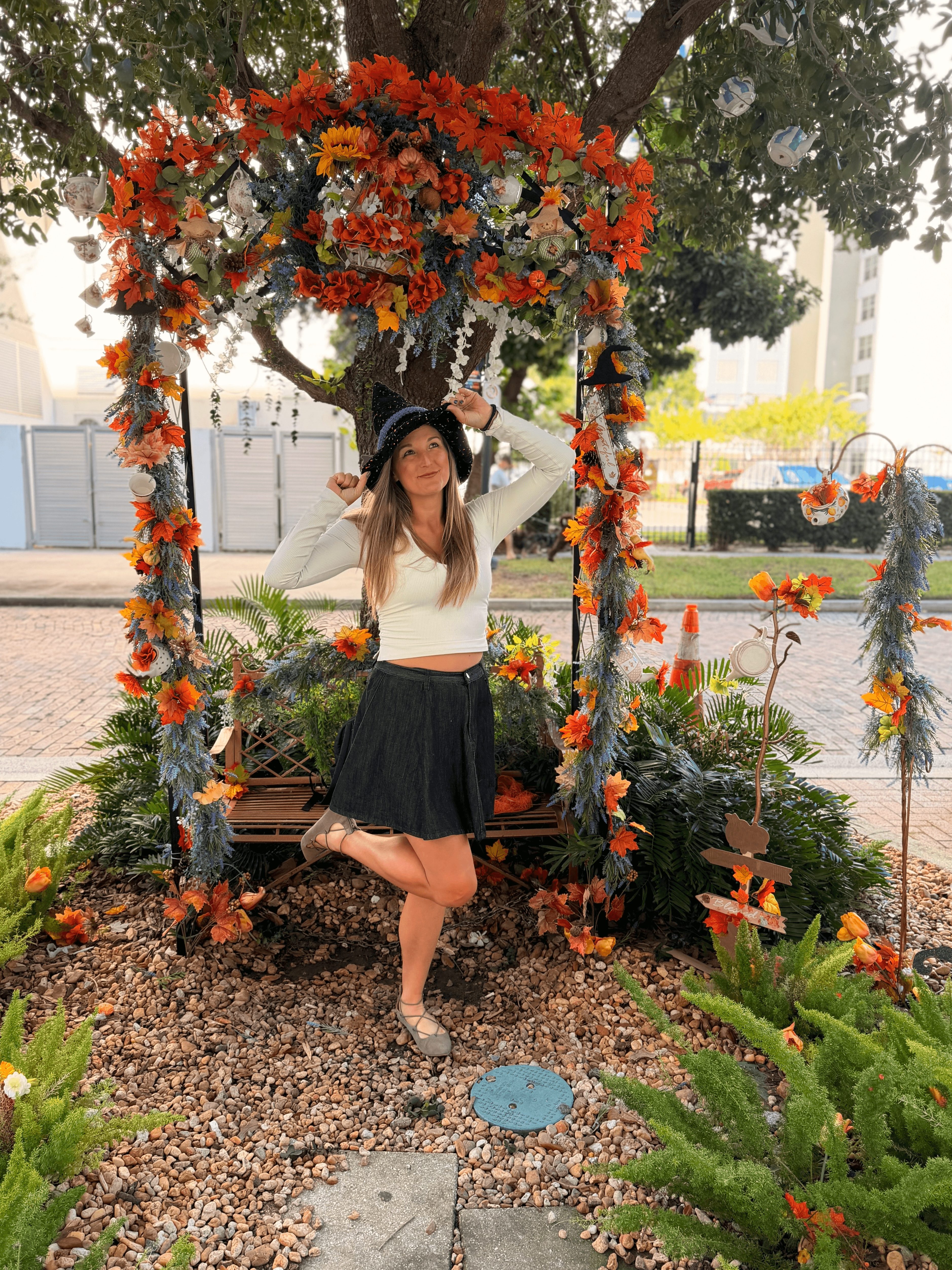 Young woman in a white top and black skirt standing on a swing decorated with orange, white, and purple flowers, outdoors.