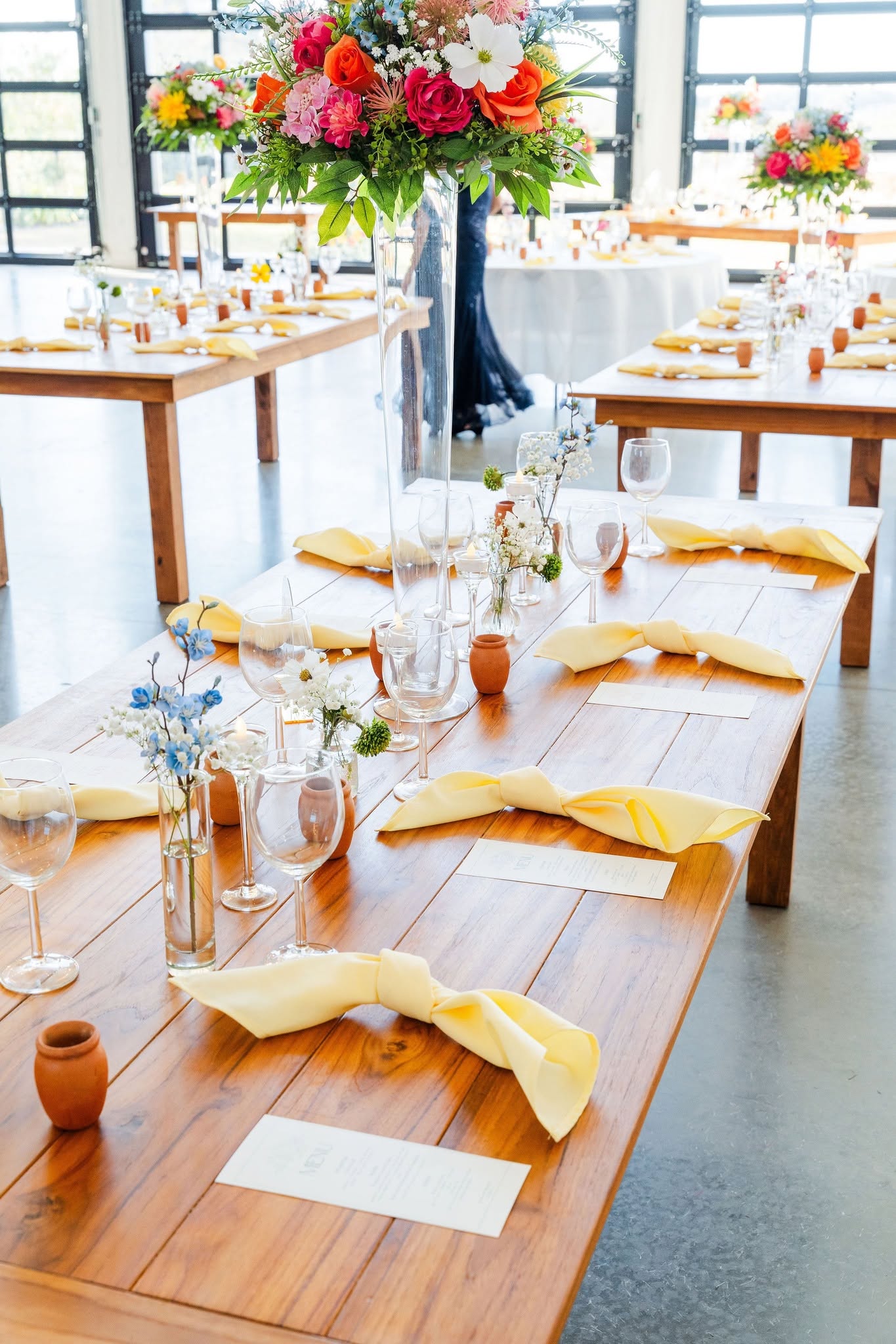 Long wooden table set with yellow napkins, glassware, and small vases, with a large floral centerpiece in a bright room.