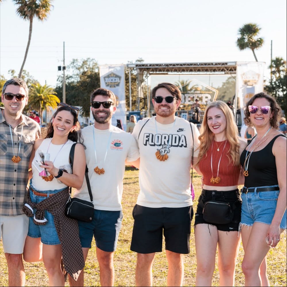Group of six people smiling outdoors at a festival, standing in front of a stage with trees and sky in background.