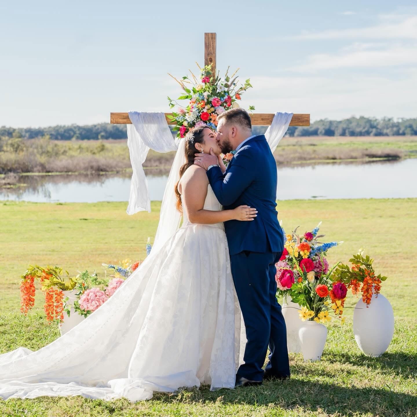 Bride and groom kiss outdoors with a cross and floral arrangements behind them, on a grassy field near water.