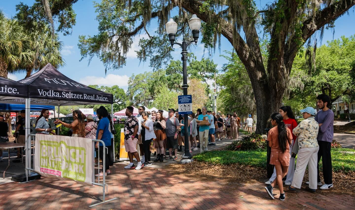 People gather in line outdoors near a tent with a sign, under a large tree with hanging moss, on a sunny day.