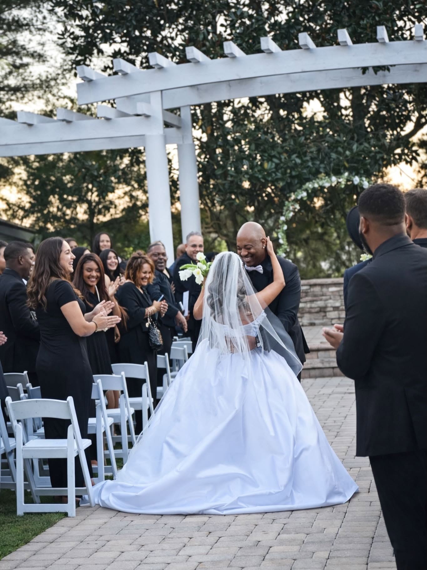 Bride and groom embrace outdoors during wedding ceremony, surrounded by guests, with white chairs and a pergola, trees in background.