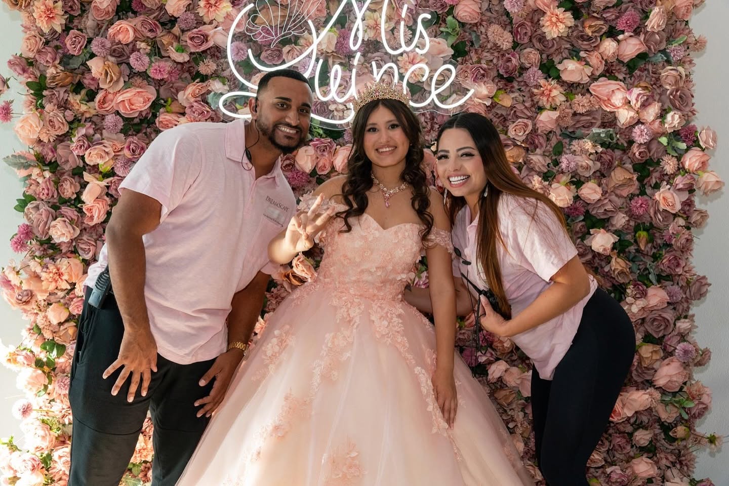 Three people smiling in front of a pink floral backdrop with a neon sign, celebrating a special occasion.