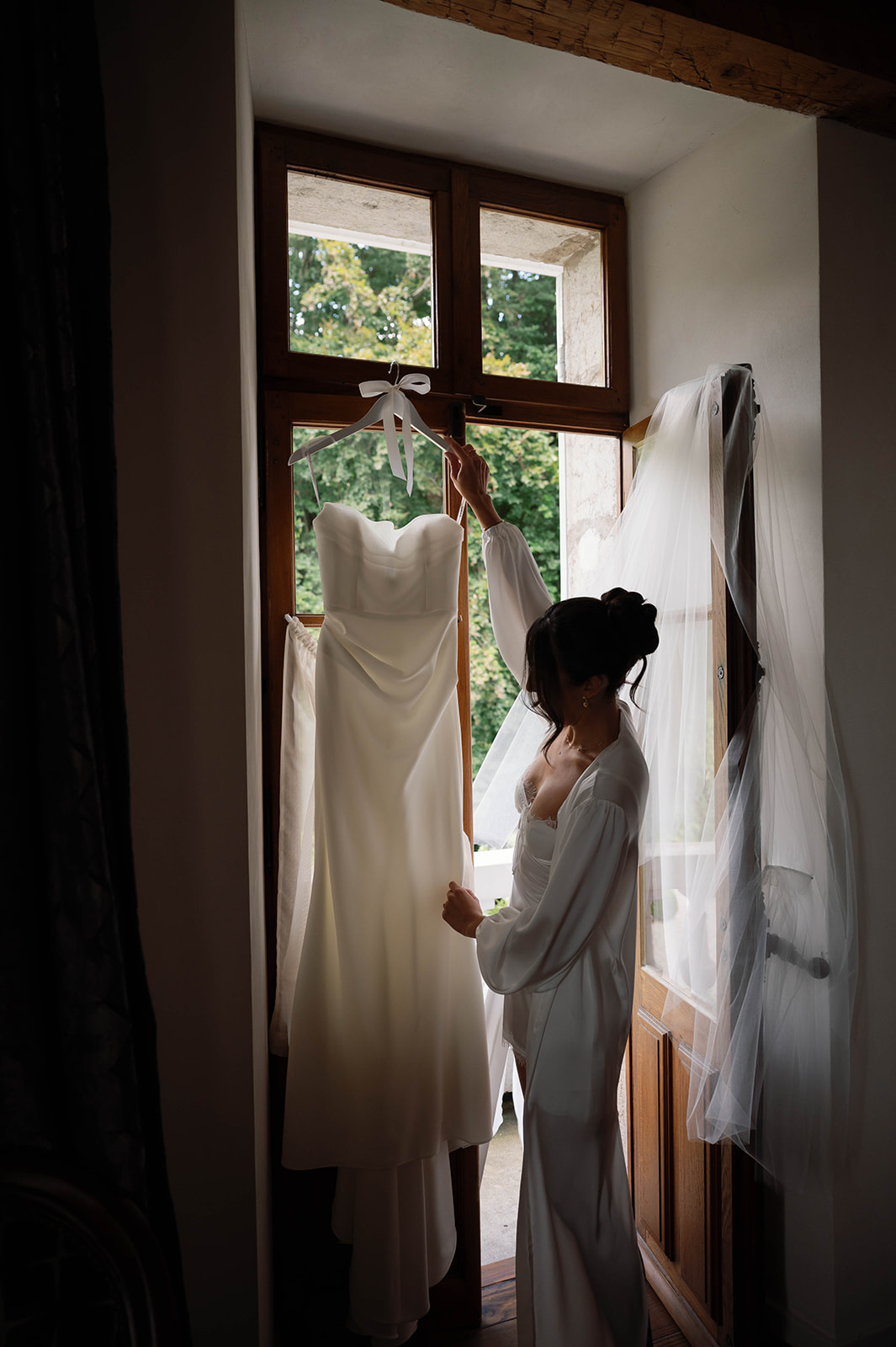 Person in white robe standing by window with wedding dress and veil hanging, sunlight streaming in, greenery outside.