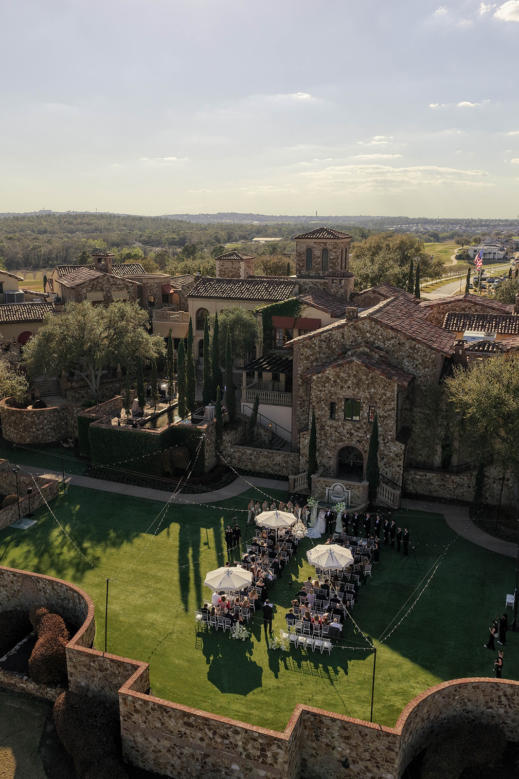 Outdoor wedding ceremony with chairs and umbrellas on a grassy area near stone buildings, under a partly cloudy sky.