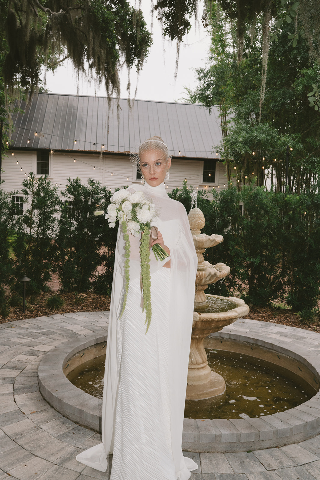 Woman in white dress holding bouquet stands near fountain with trees and building in background.
