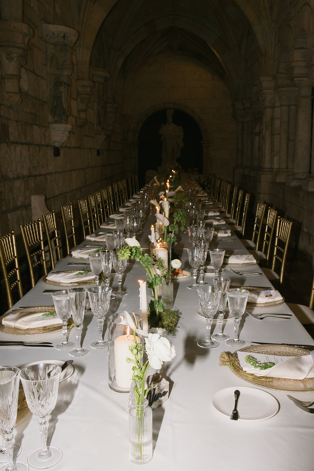 Long banquet table with white tablecloth, floral centerpiece, glassware, and place settings in a stone archway hall.
