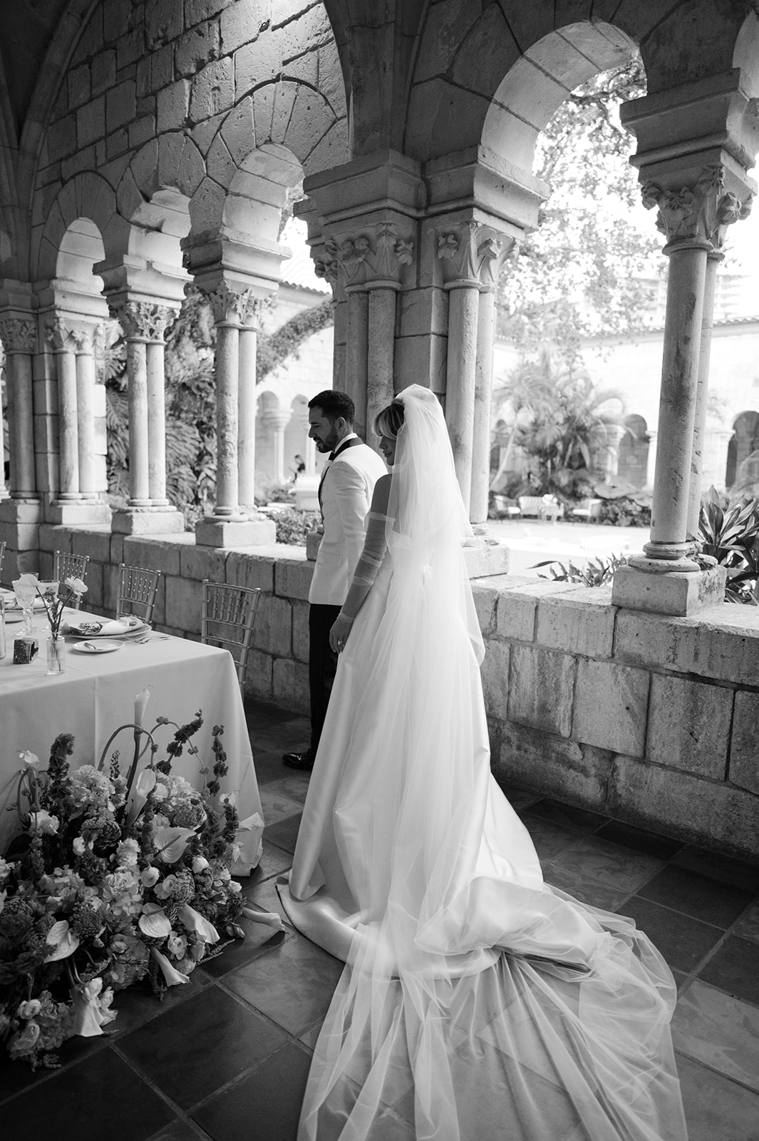 Bride and groom stand together under arches, with floral arrangements nearby, in a black and white wedding scene.