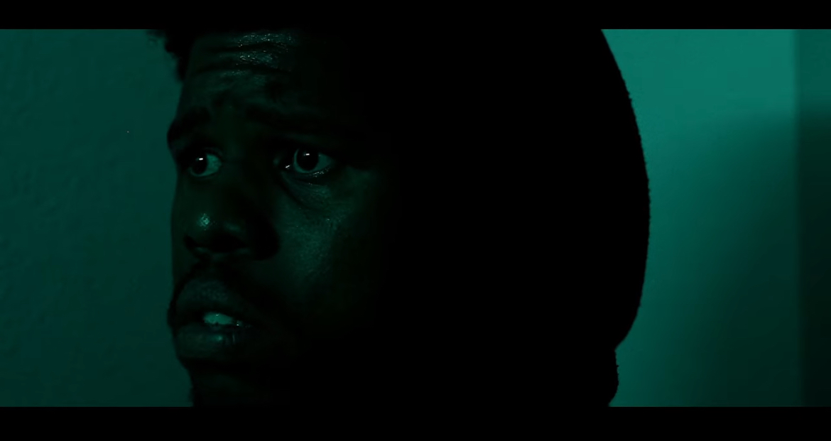 Close-up of a person's face in dark lighting, with a serious expression, looking to the side.