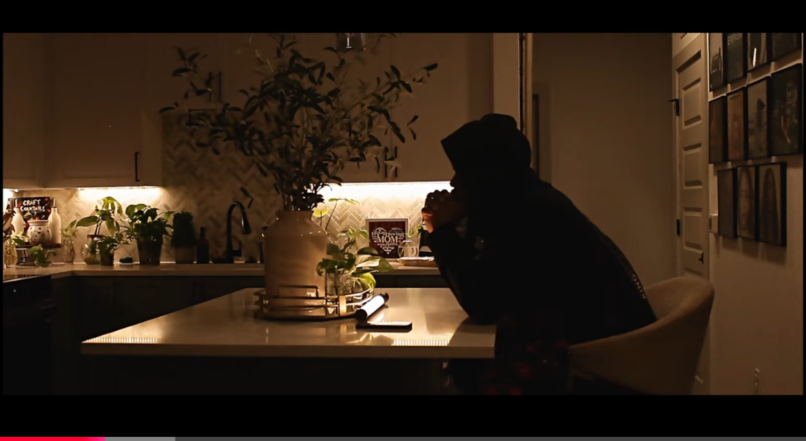 Person sitting at a table in a dimly lit room, drinking from a cup, with plants and pictures on the wall.