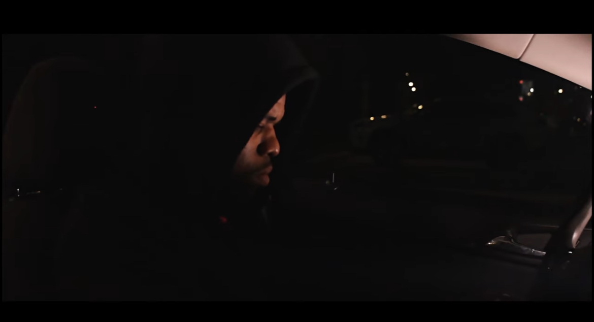 Person wearing a hooded jacket sitting in a car at night, with city lights visible outside.
