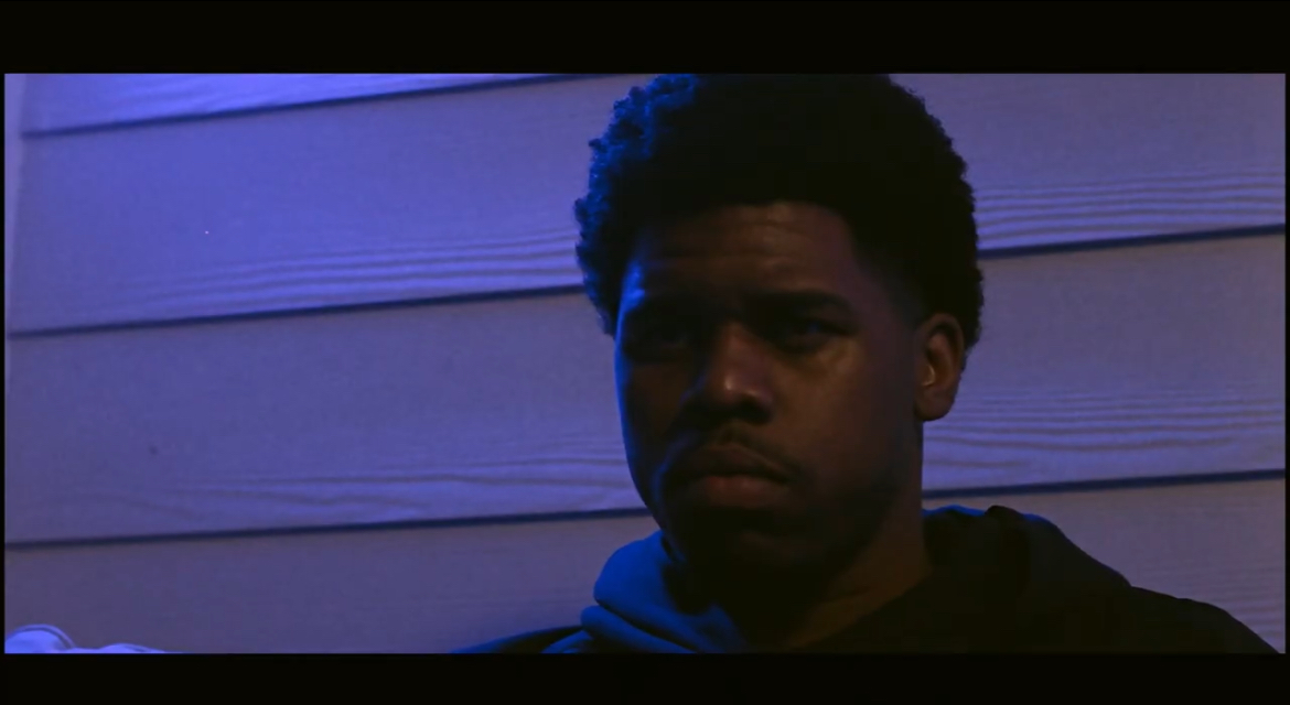 Young man with dark hair and serious expression, standing against a light-colored wall, illuminated by blue light.