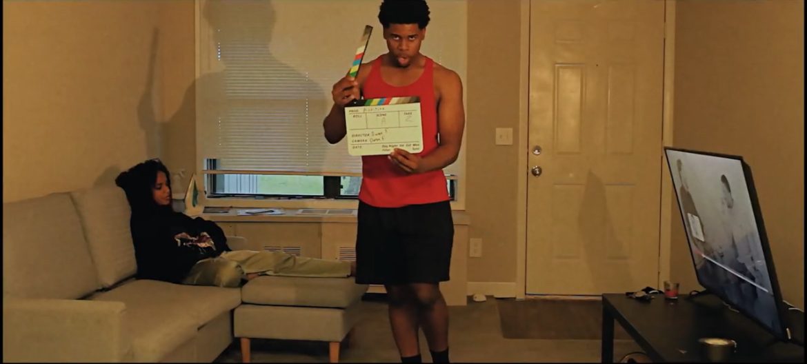 Person holding a clapperboard standing in a living room with a woman sitting on a sofa, TV on a stand, and a door in the background.