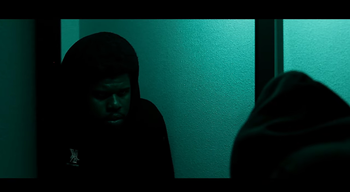 Two people sitting in a dimly lit room with frosted glass windows, one facing forward and the other with their back turned.
