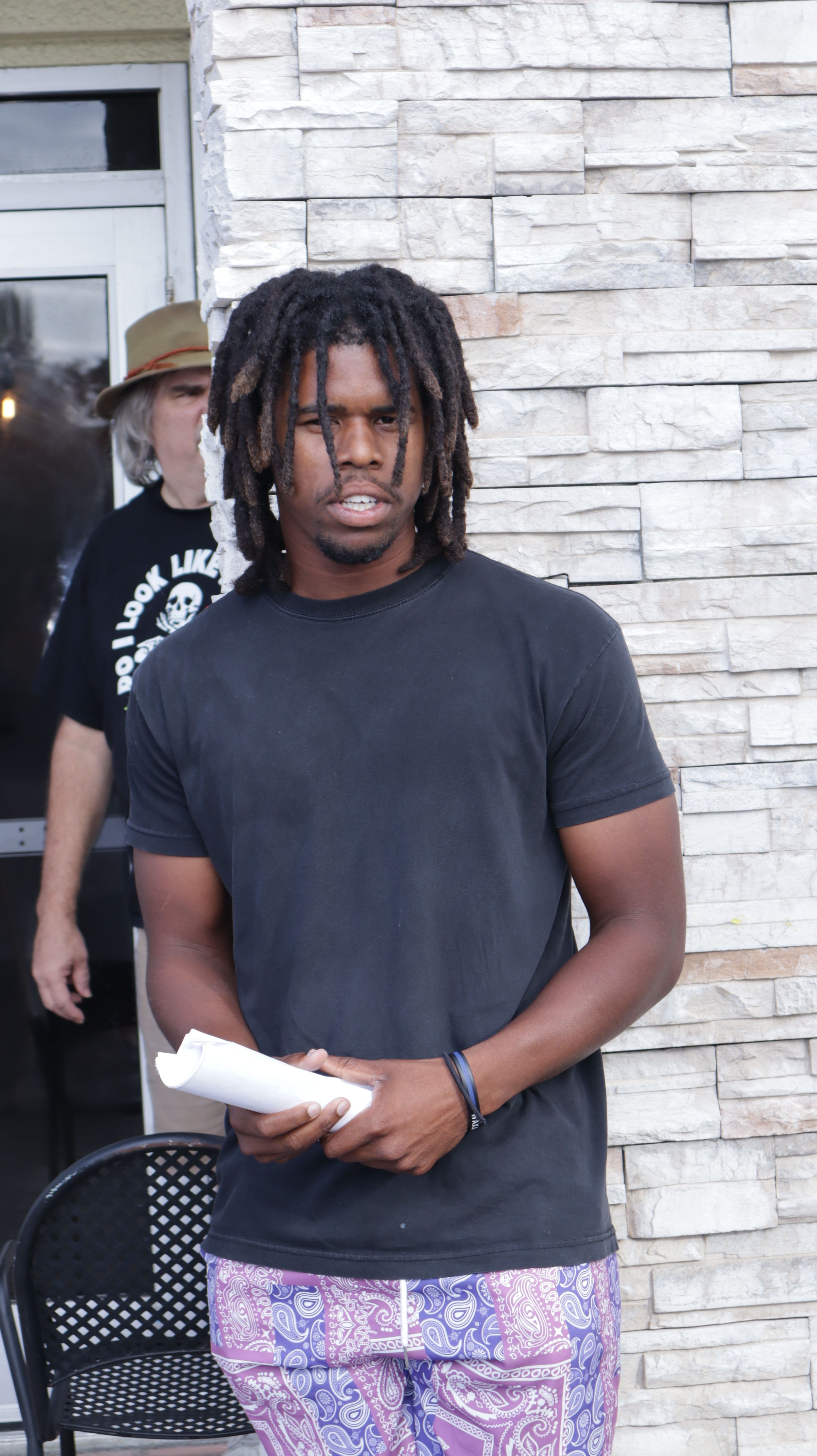 Person with dreadlocks holding a paper, standing against a white brick wall, wearing a black t-shirt and patterned pants.