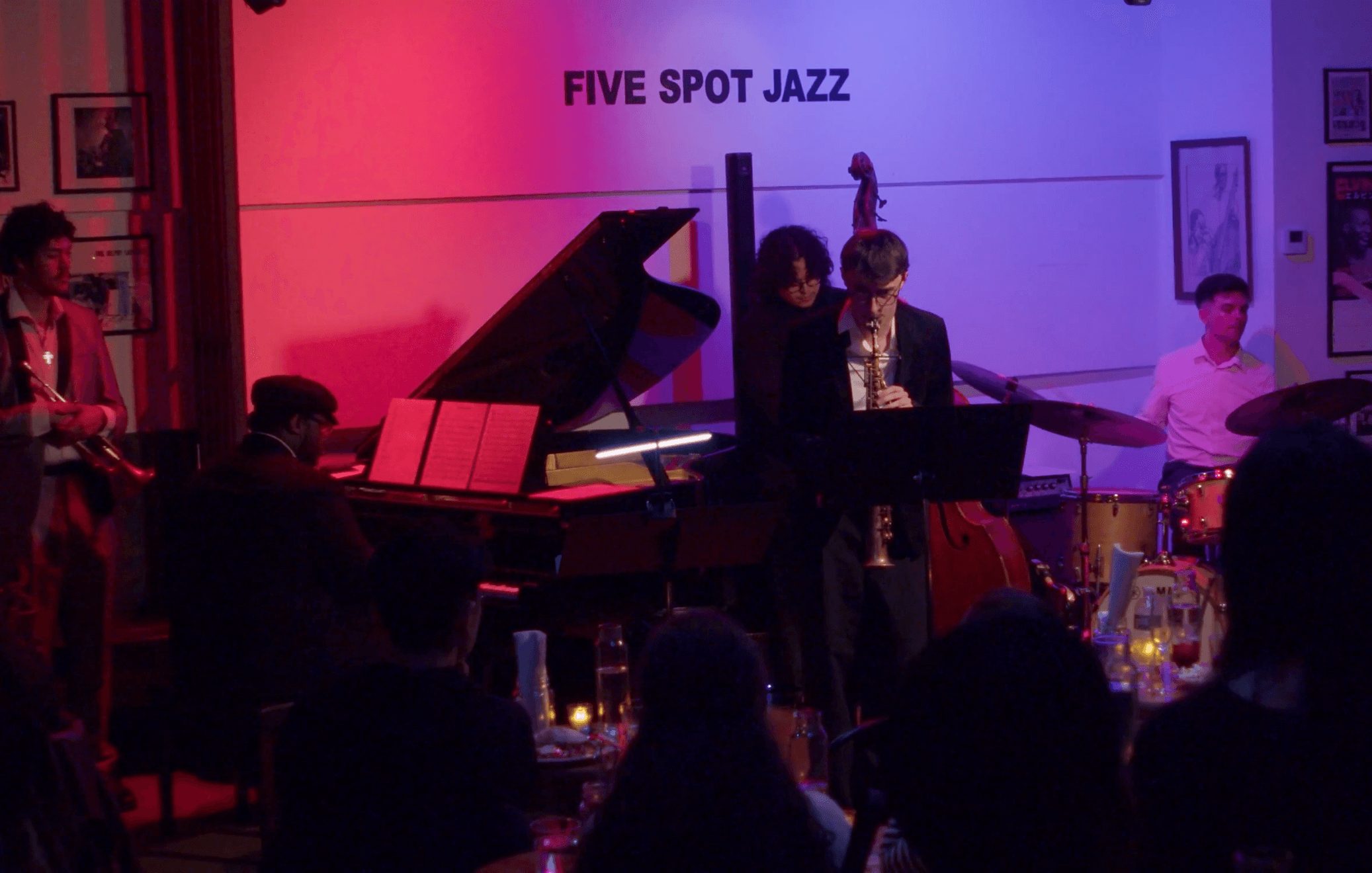 Jazz band performing with piano, drums, and a singer in a dimly lit venue, audience seated at tables, sign reads 'FIVE SPOT JAZZ'.