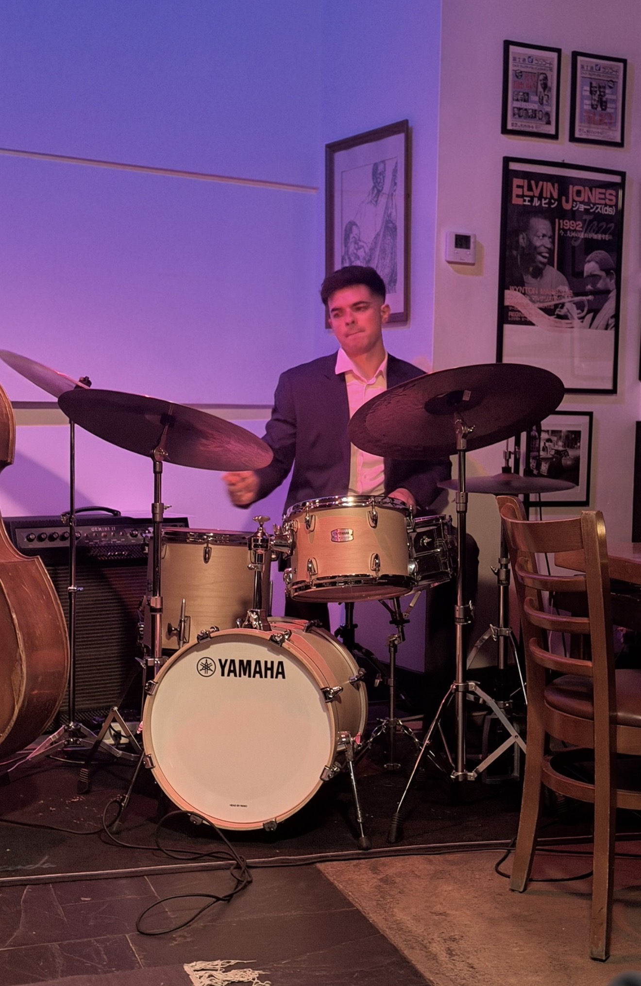 Young man in a suit playing a Yamaha drum set indoors, with framed posters on the wall behind him.