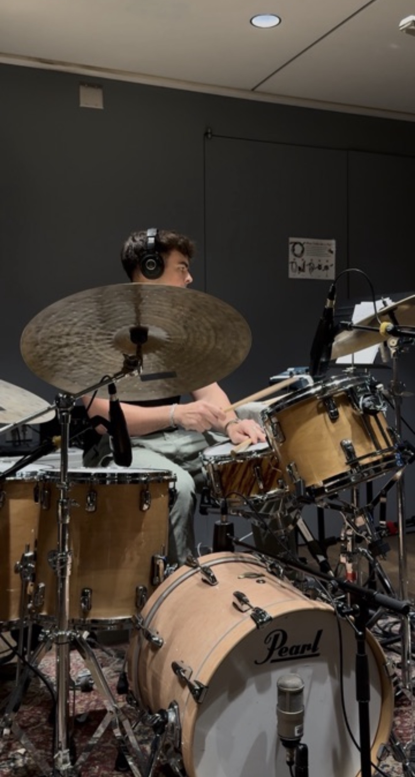 Person playing drums in a music studio, wearing headphones, surrounded by drum equipment and cymbals.