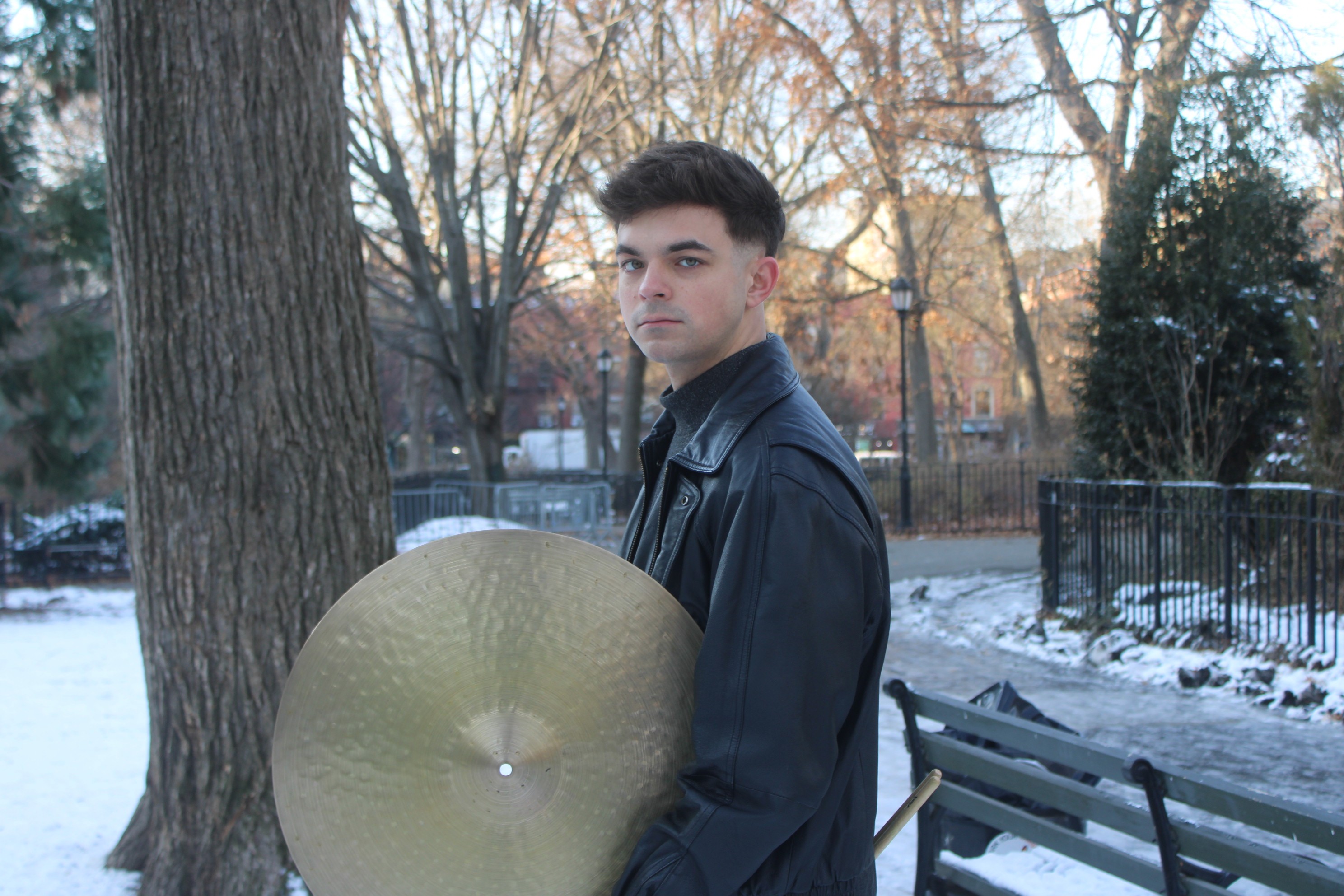 Young man standing outdoors in winter, wearing a dark jacket, with trees and a park bench in the background.