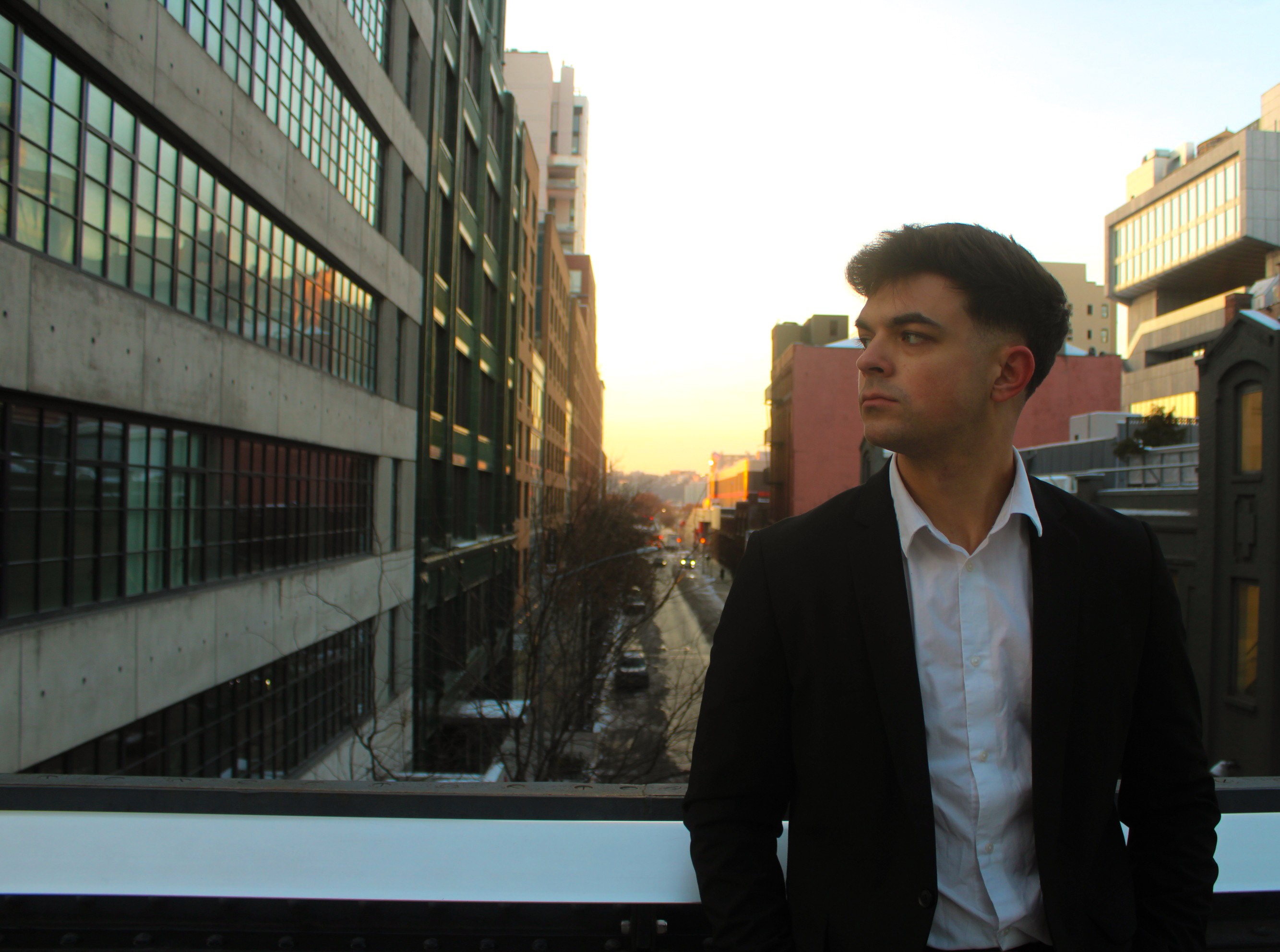 Young man in formal attire stands on a bridge in an urban setting during sunset, with buildings and a canal in the background.