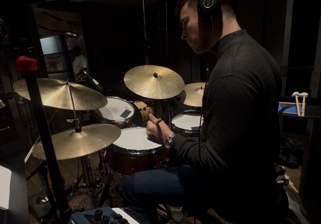 Person wearing headphones playing drums in a recording studio, surrounded by cymbals and drum equipment.