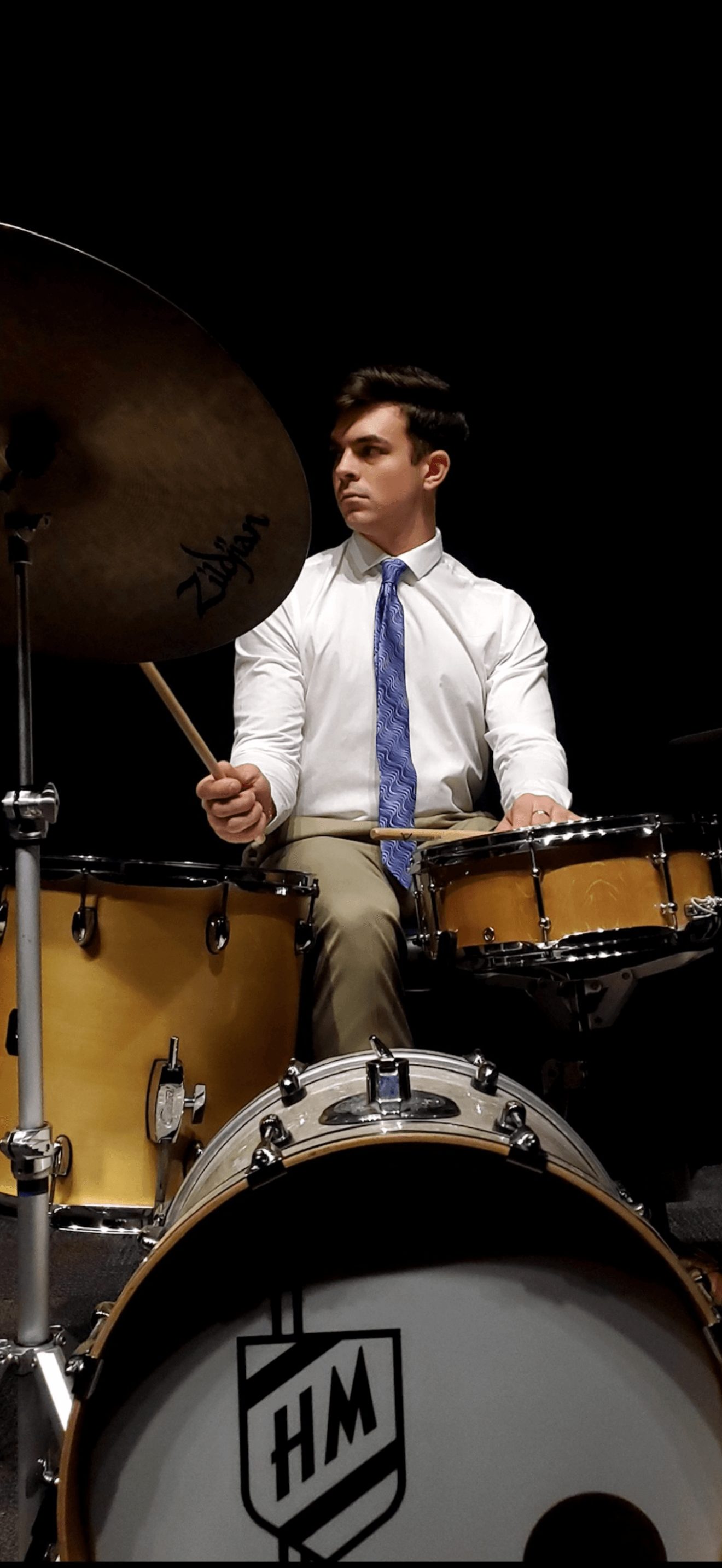 Young man playing drums, wearing white shirt and blue tie, dark background, focused expression.