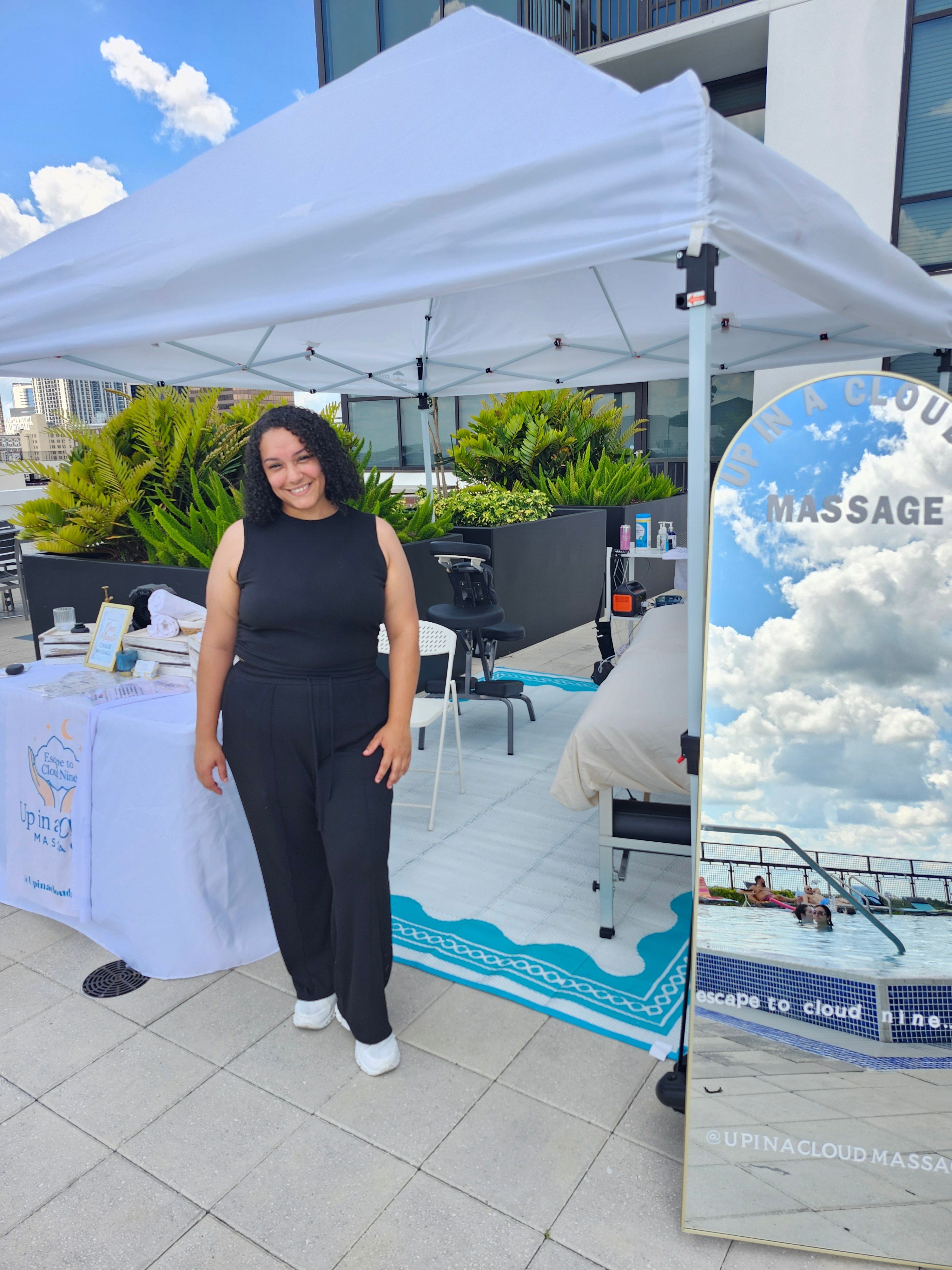 Woman in black dress standing next to a display booth outdoors, with a table and promotional materials, under a white canopy.
