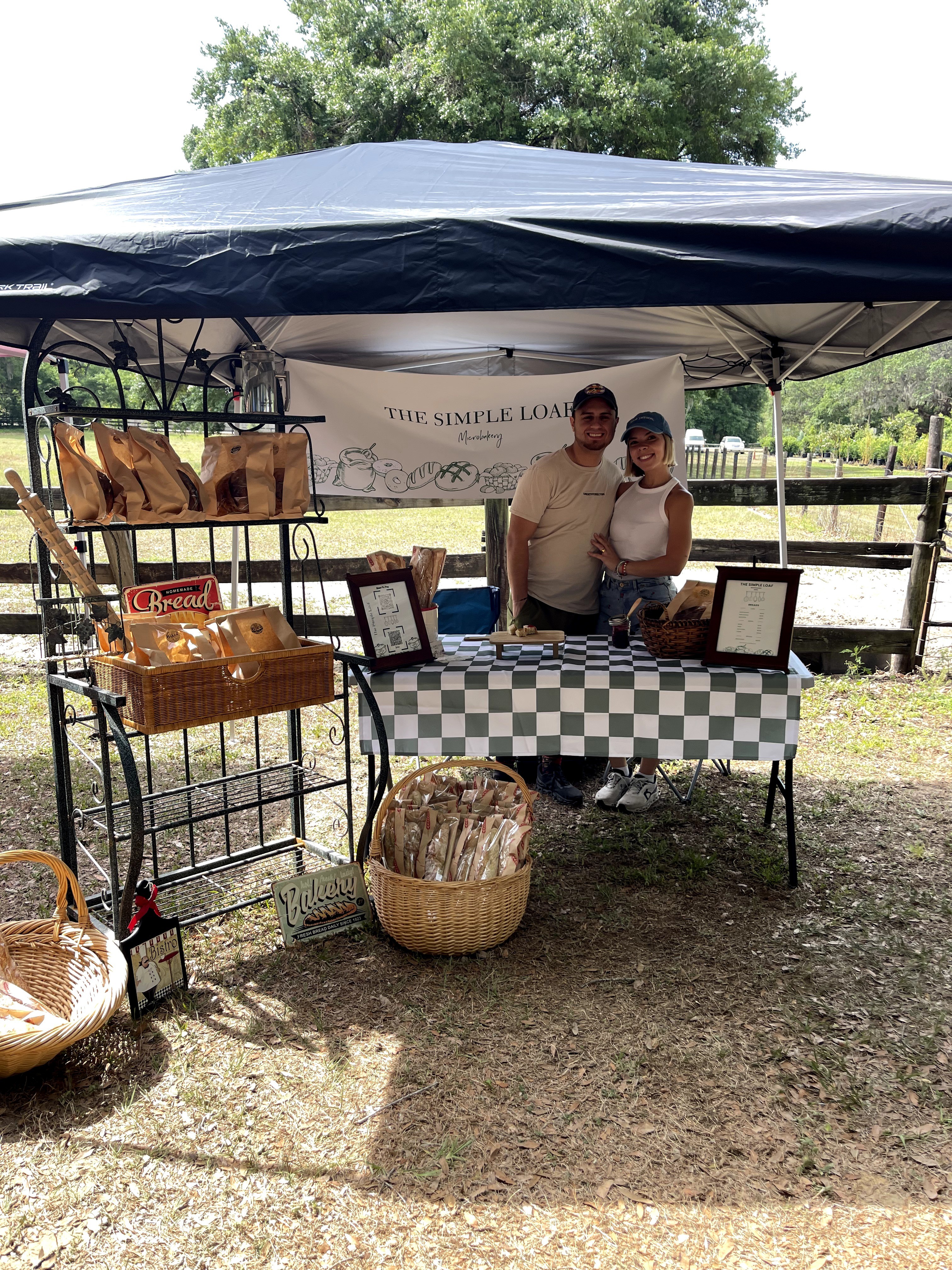 Two people stand behind a table with framed items, under a canopy, outdoors with trees and a fence in background.