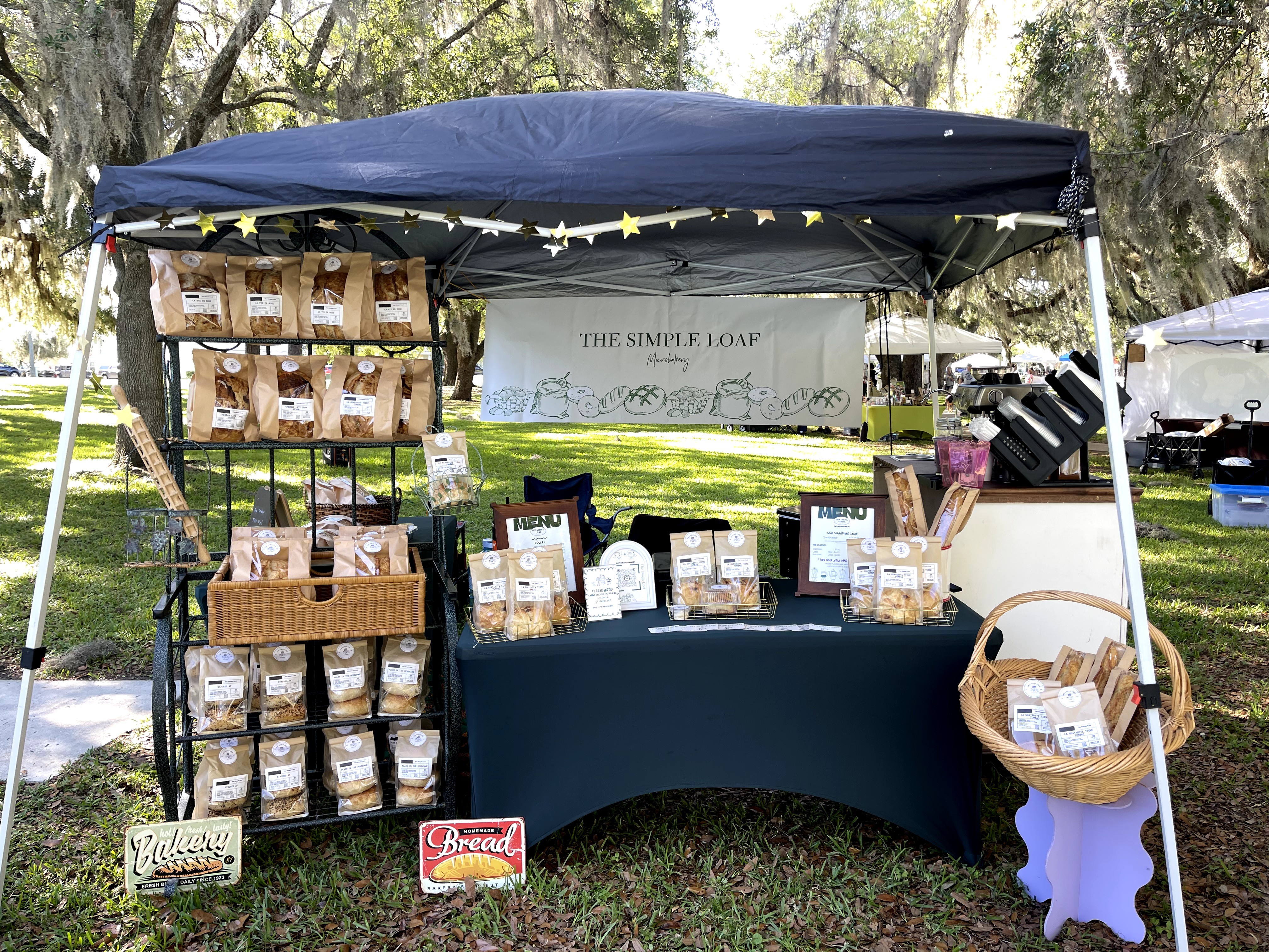 Outdoor market stall with bags and products under a blue canopy, trees in background, table with display items, and baskets.