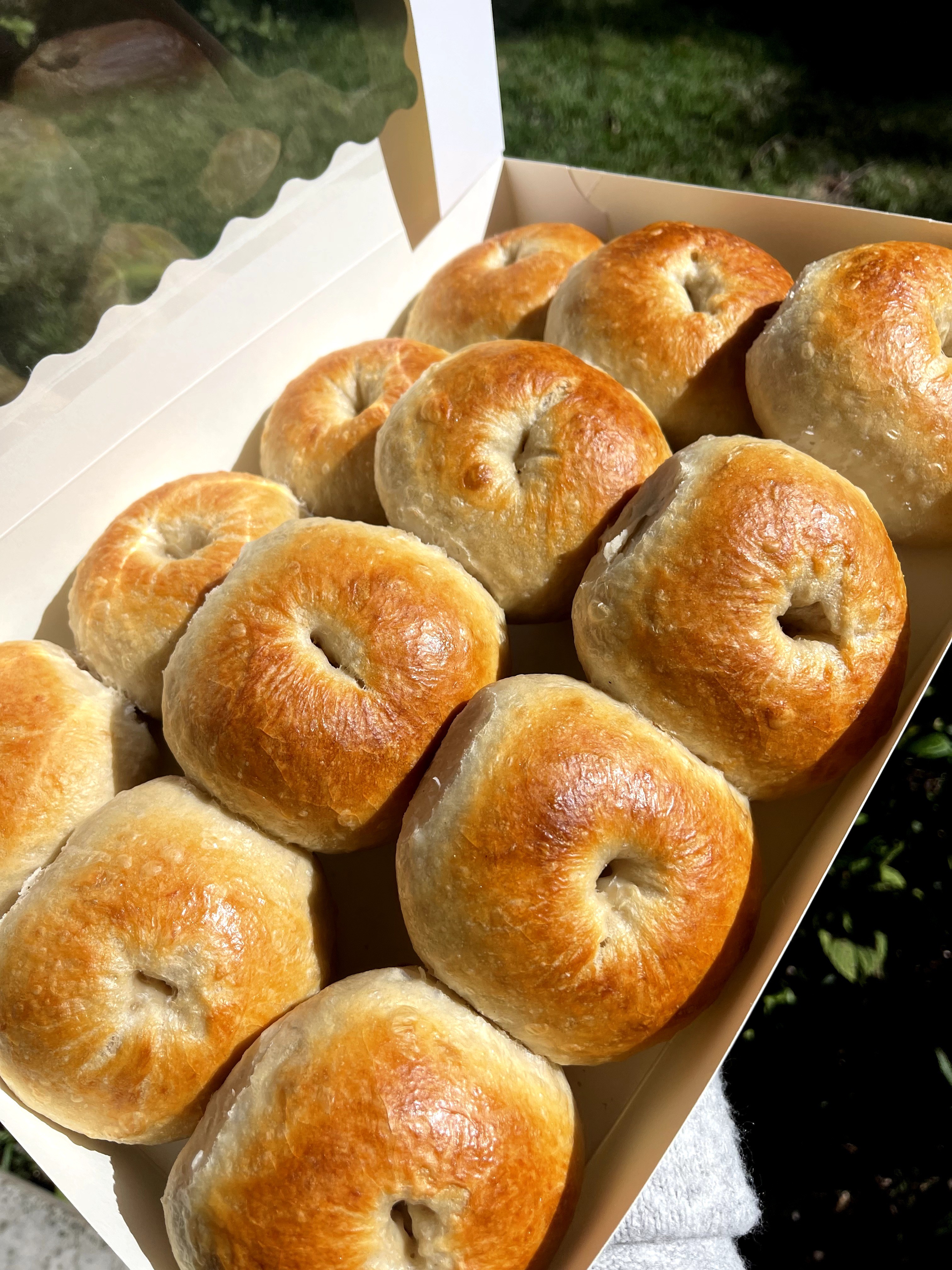 Box of golden-brown baked bread rolls on a white tray outdoors, with green grass in the background.