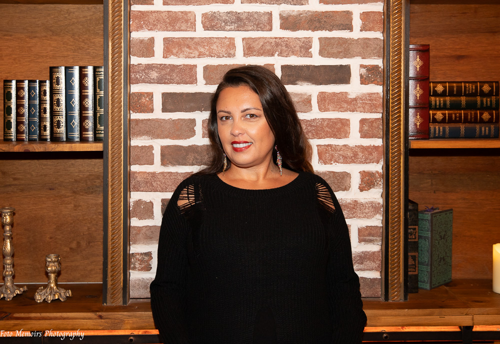 Woman with dark hair and red lipstick standing in front of a brick wall, flanked by wooden bookshelves.