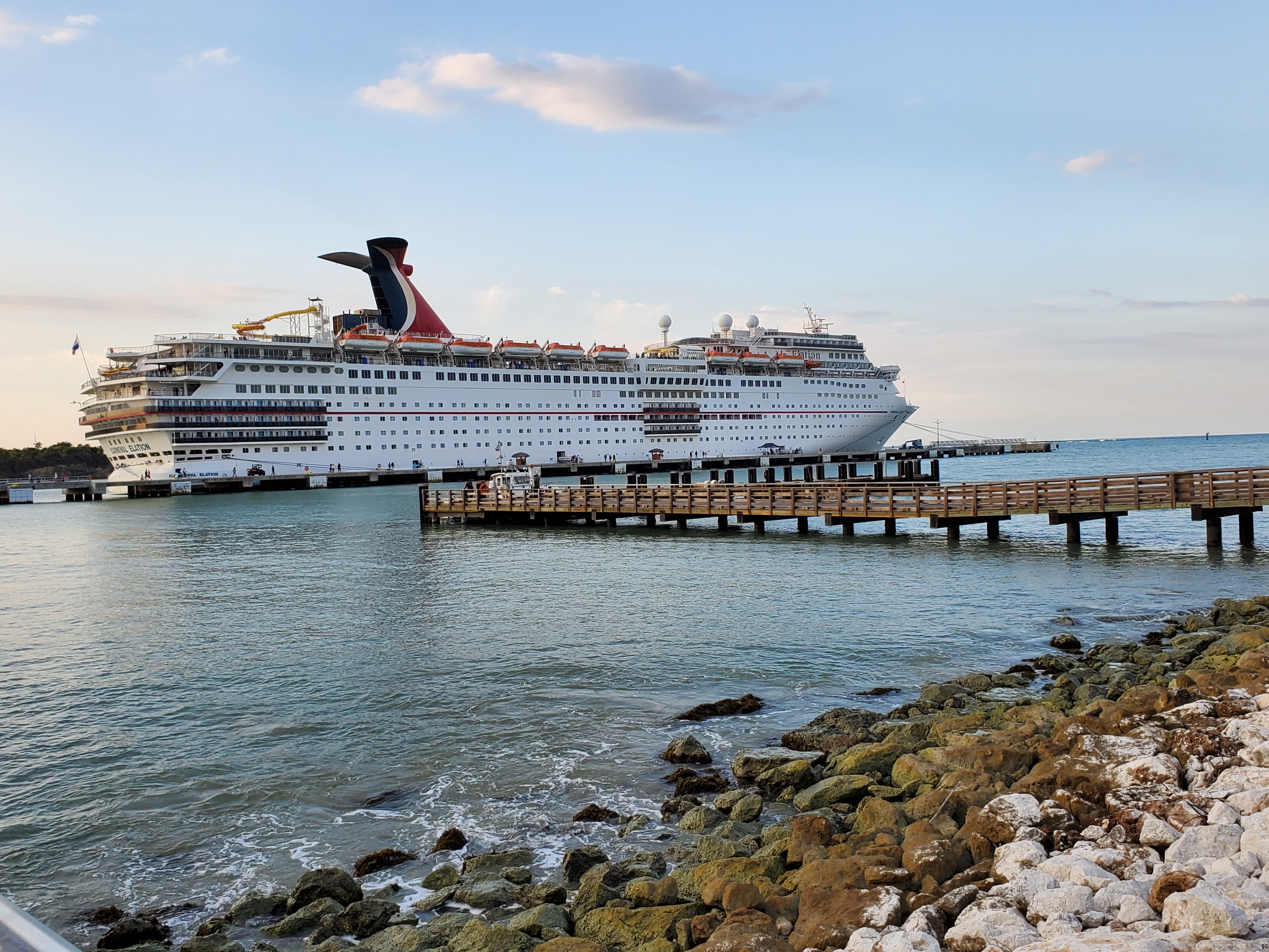 Large cruise ship docked at port with a pier extending into water, rocky shoreline in foreground, sky with clouds above.
