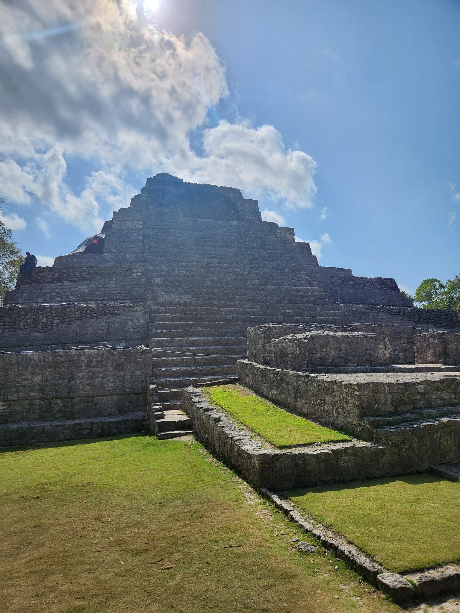 Ancient stepped pyramid structure with stone stairs, surrounded by green grass and a partly cloudy sky.