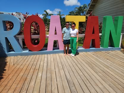 Two people stand in front of large colorful letters spelling 'ROATAN' on a wooden deck, with trees and a building in the background.