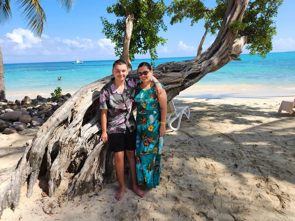 Two women stand on a sandy beach near a large tree with the ocean in the background, smiling at the camera.