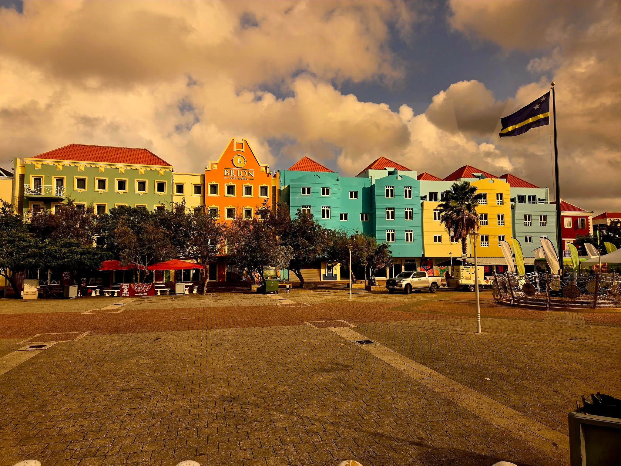 Colorful buildings with a flag on a pole, trees, and a park area under a cloudy sky.