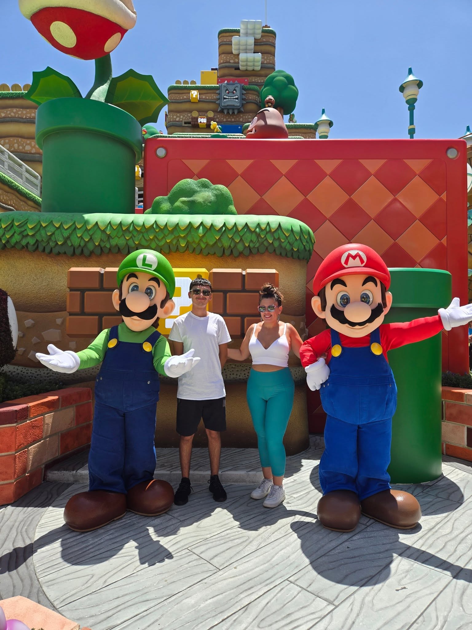Three people stand between two large Mario and Luigi statues in a colorful theme park setting, with game elements in the background.