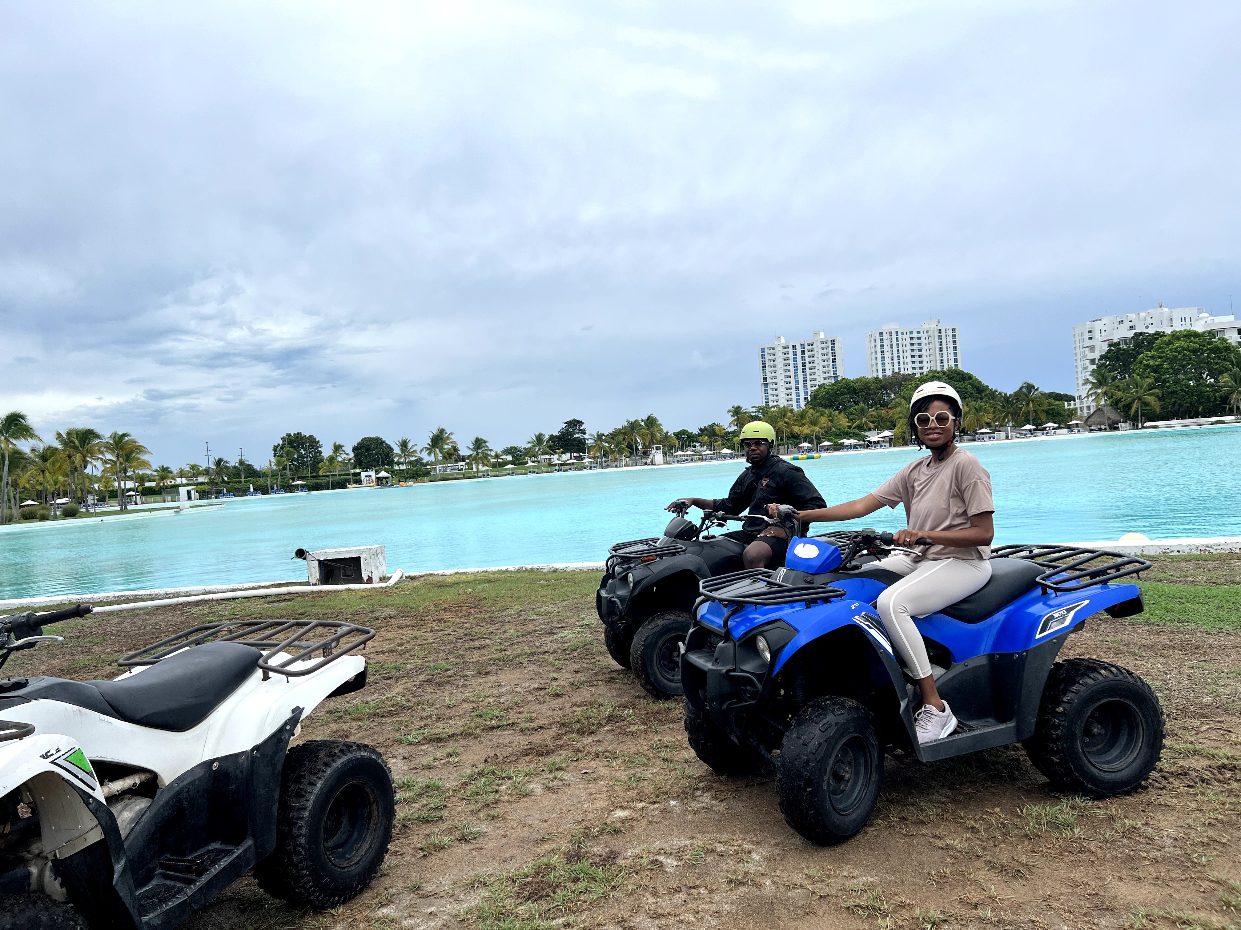 Two people sitting on blue and black all-terrain vehicles near a body of water with buildings in the background.
