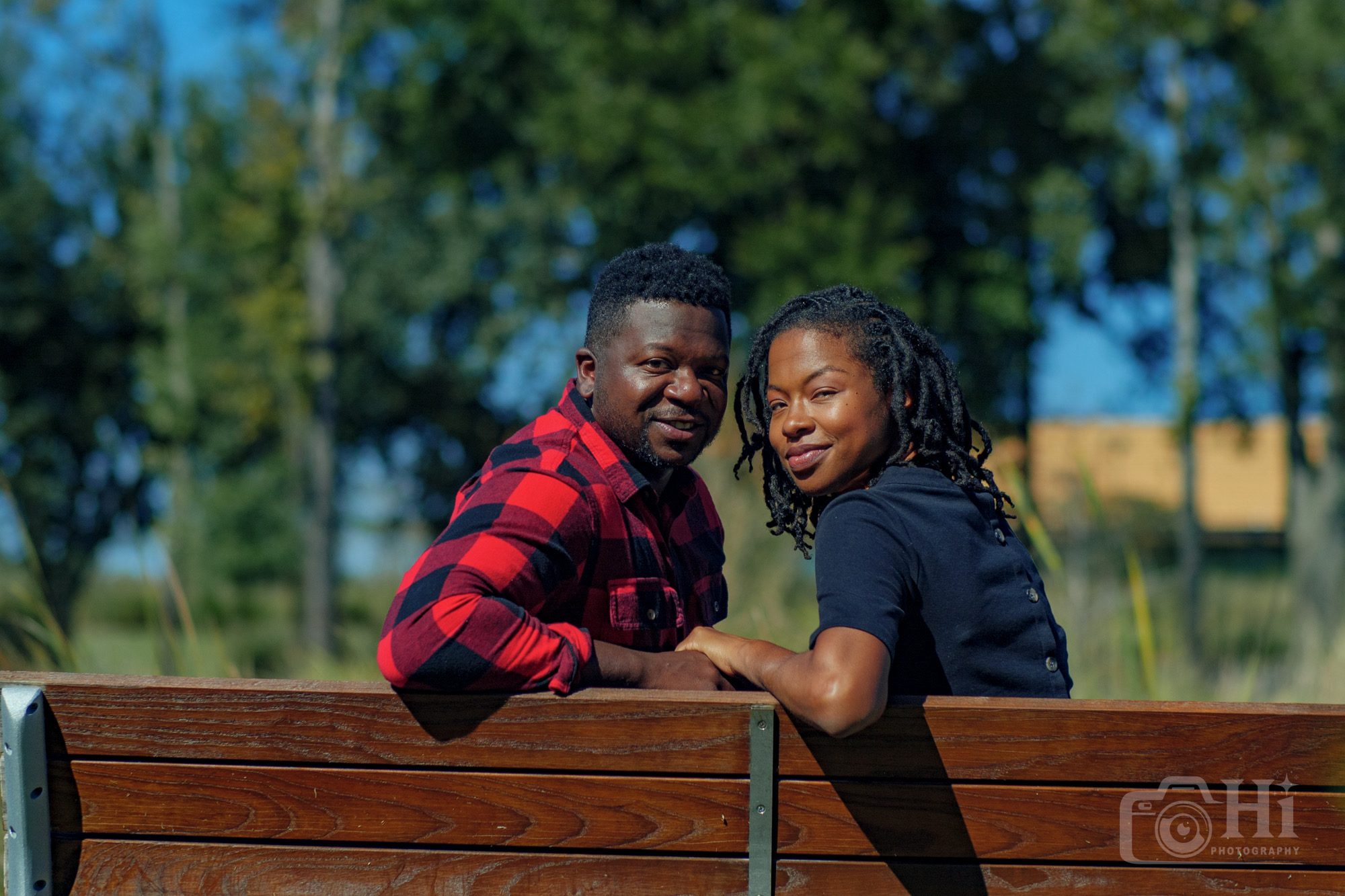 Two people sitting on a wooden bench outdoors, smiling, with trees and a blue sky in the background.