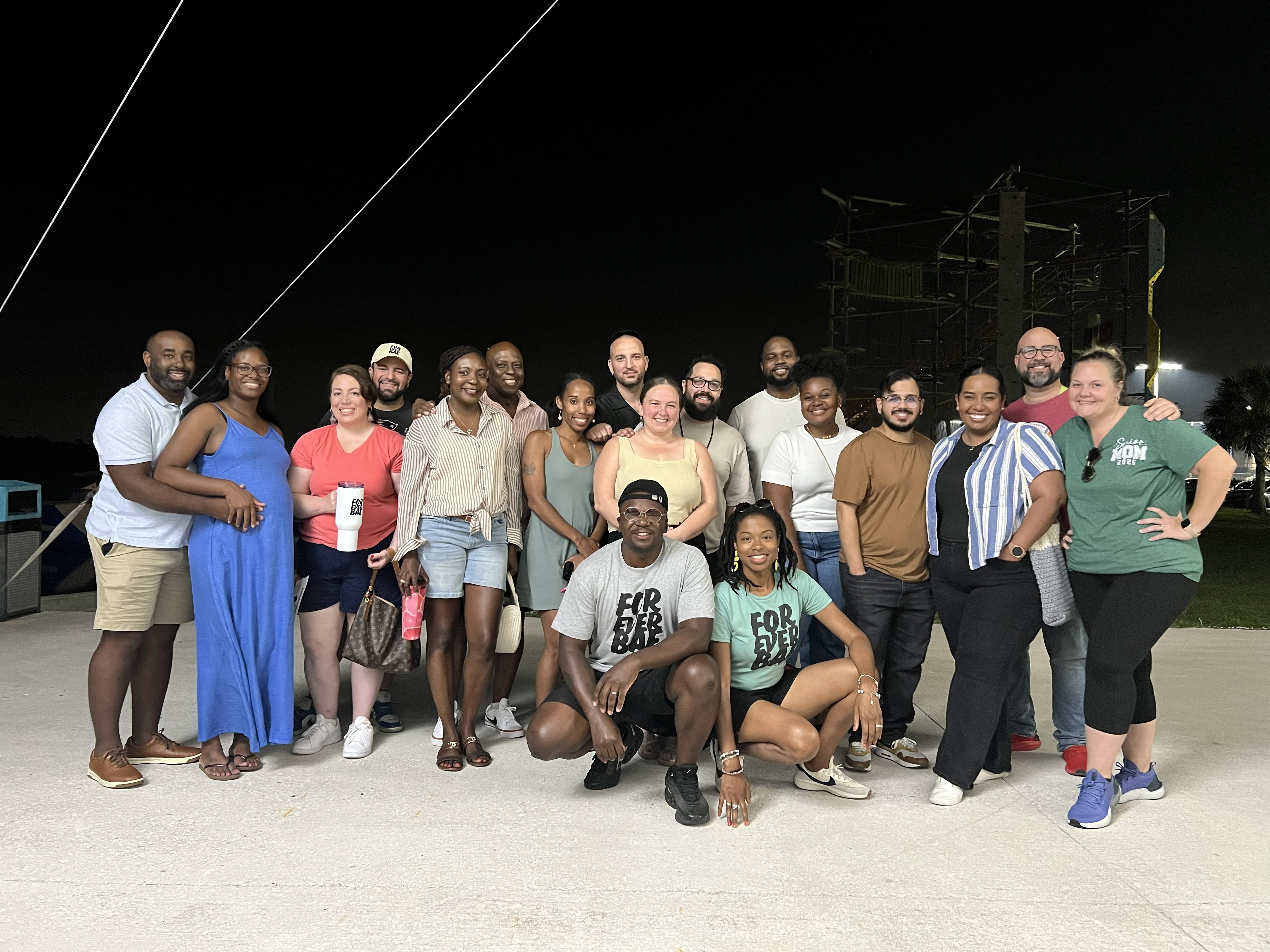 Group of people standing outdoors at night, smiling, with power lines and structures in the background.