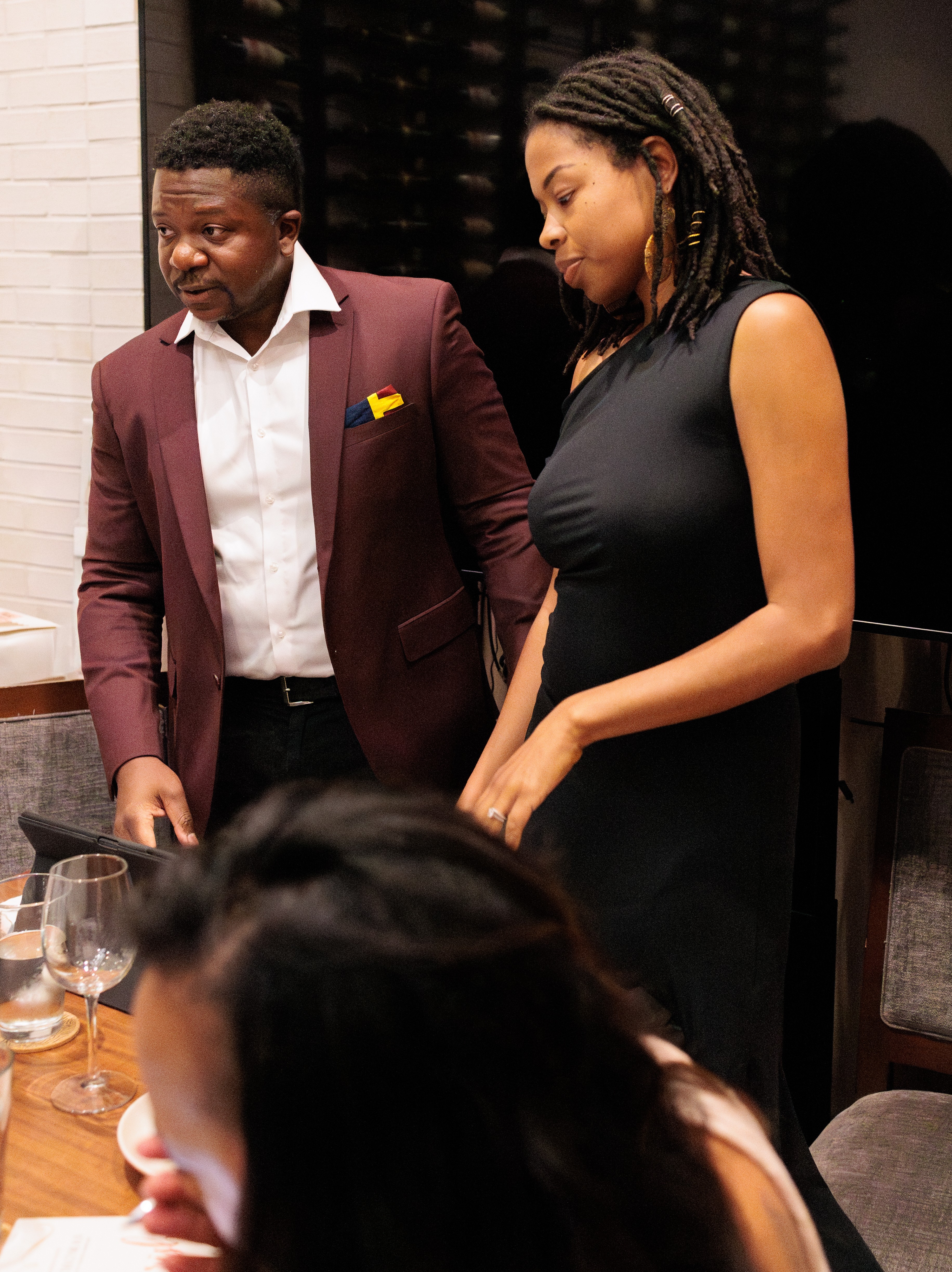 Two people standing and talking at a table, with a third person partially visible in the foreground.